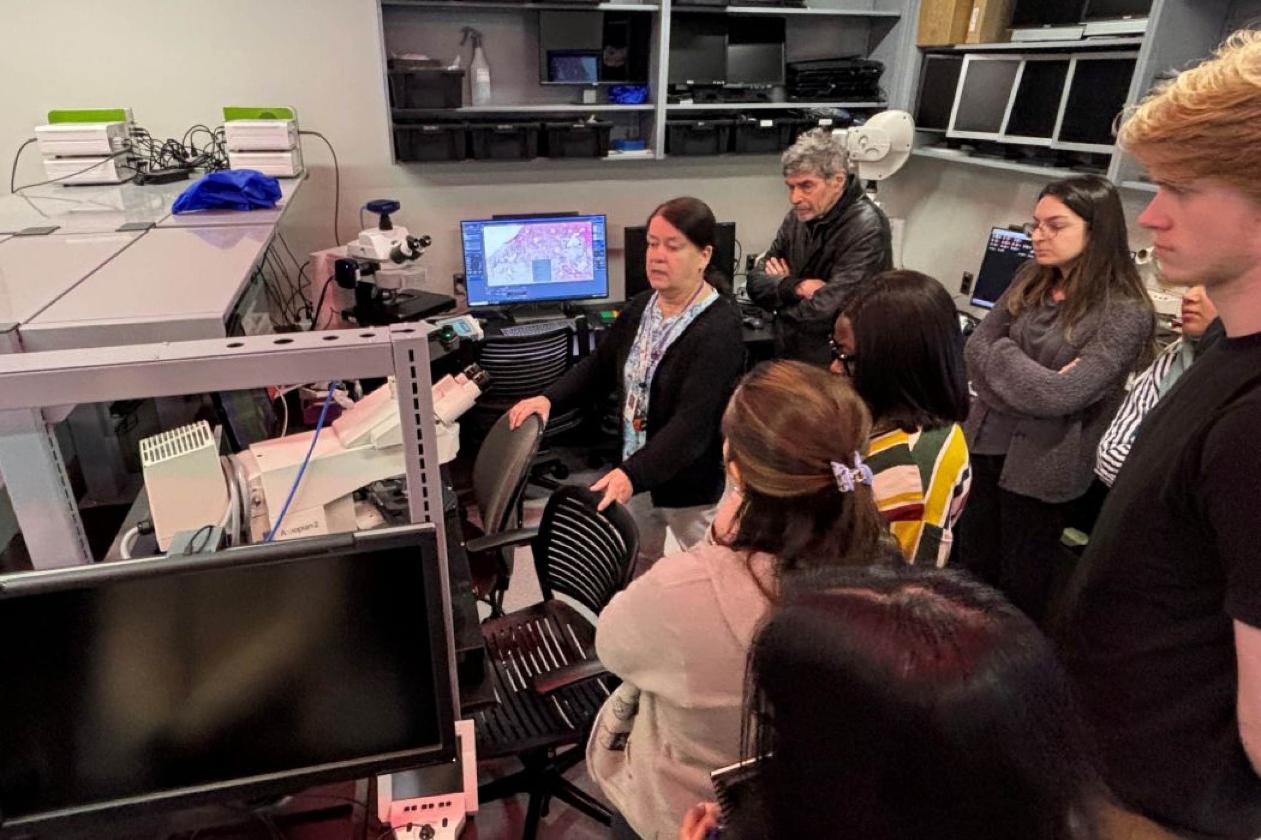 Seven people stand around Dr. Sabine Mai. There are microscopes and computer monitors in the room.