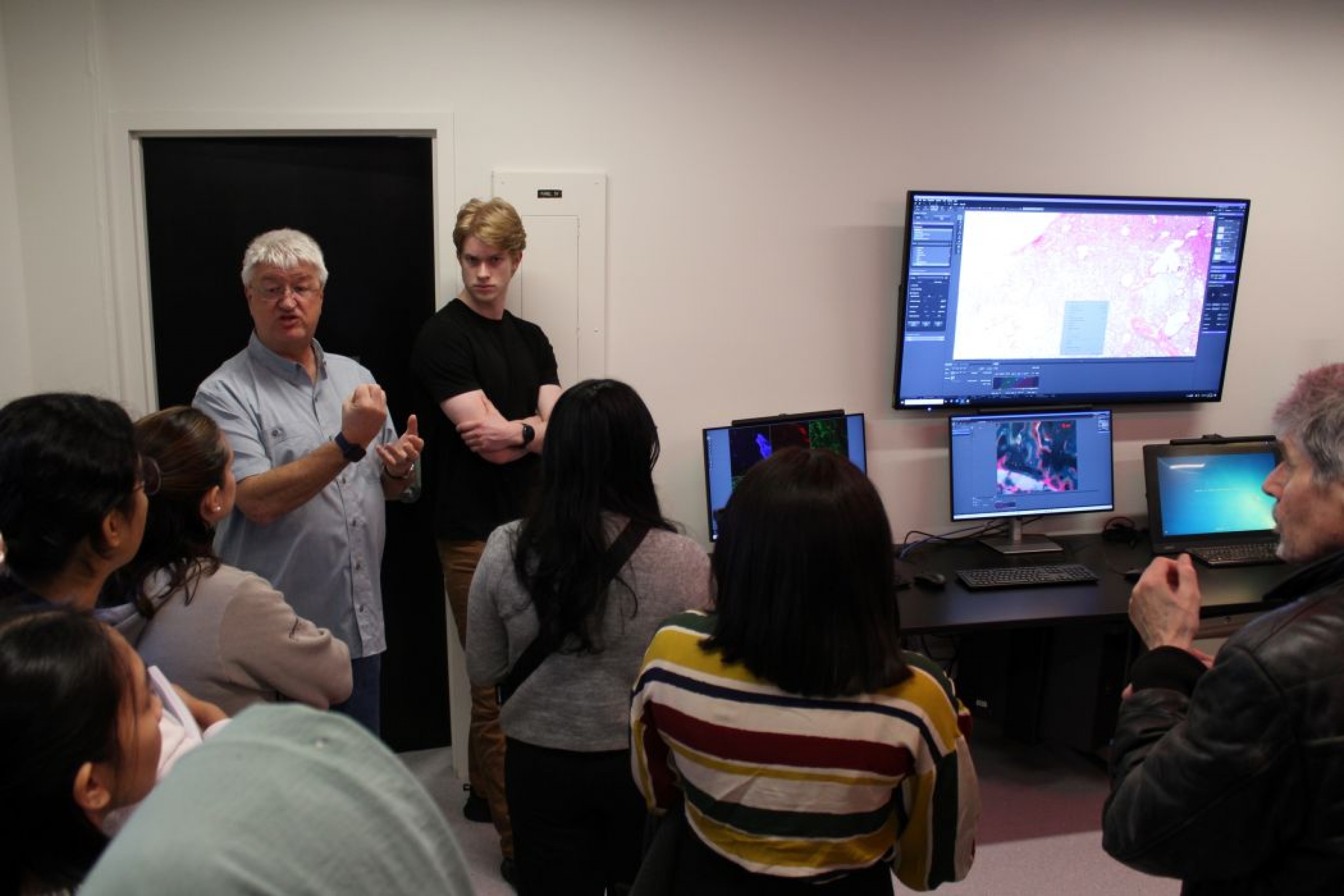 Seven people gather around Darryl Dyck. Three computer monitors and a large screen feature colourful graphics.
