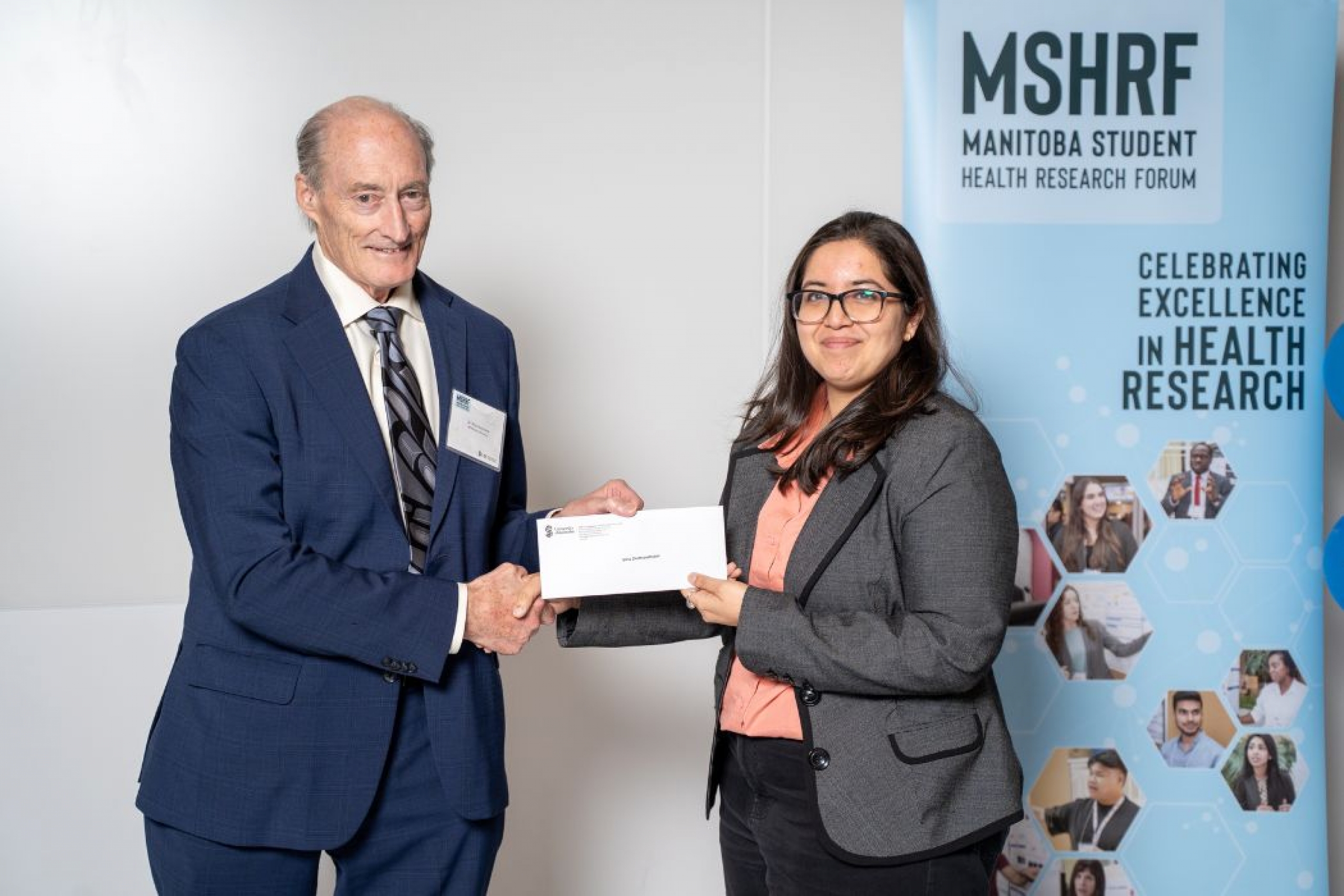 A person presenting an award to a student, behind them there's a banner that says MSHRF - Manitoba Student Health Research Forum, Celebrating Excellence in health research.