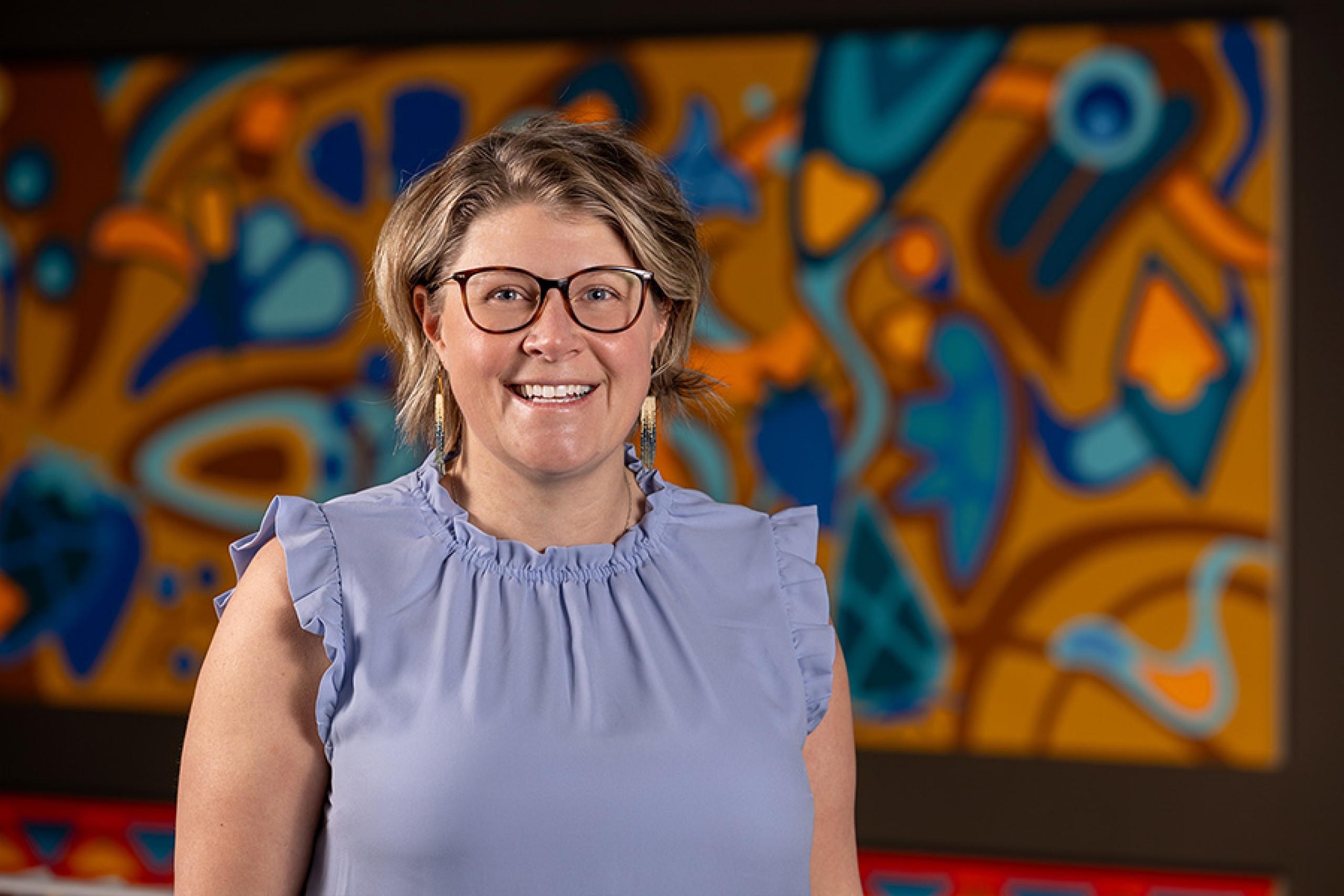A woman in a periwinkle shirt smiles in a photo. There is an oversized colourful piece of artwork as the background.