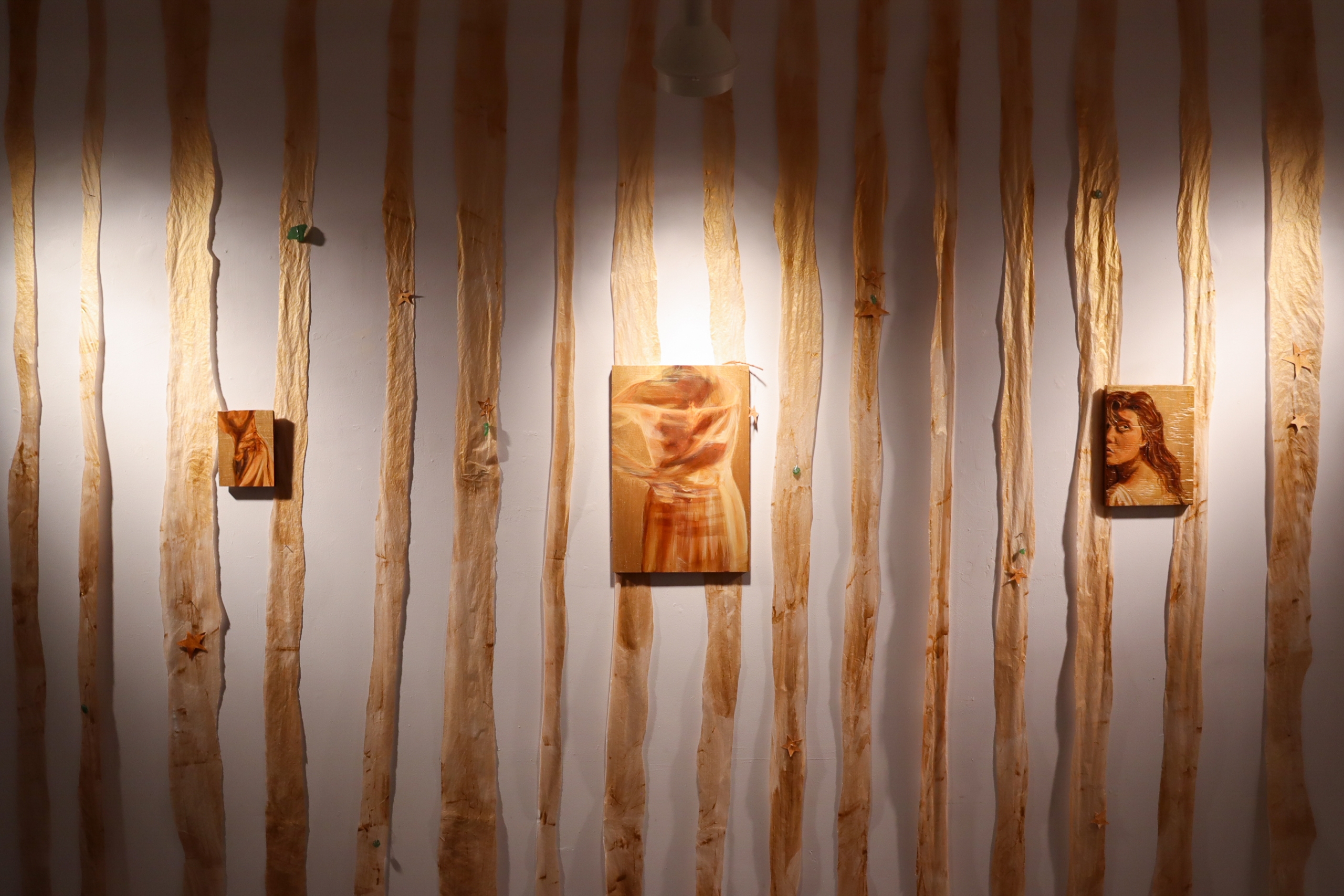 Three small paintings hang among long vertical strips of stained fabric pinned to a white gallery wall under spotlighting.
