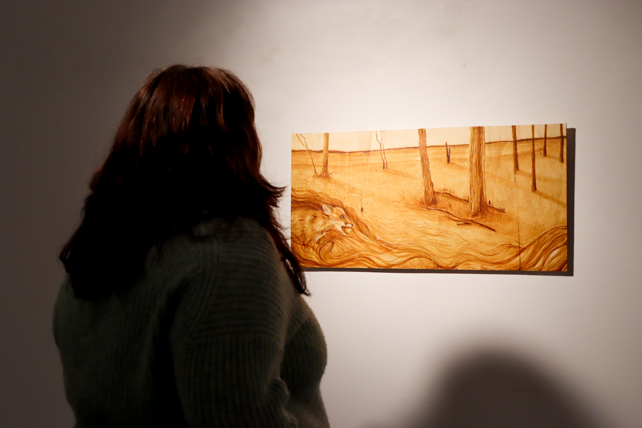 A visitor stands facing a wide rectangular panel depicting a sparse landscape with trees in warm brown tones on a white gallery wall.