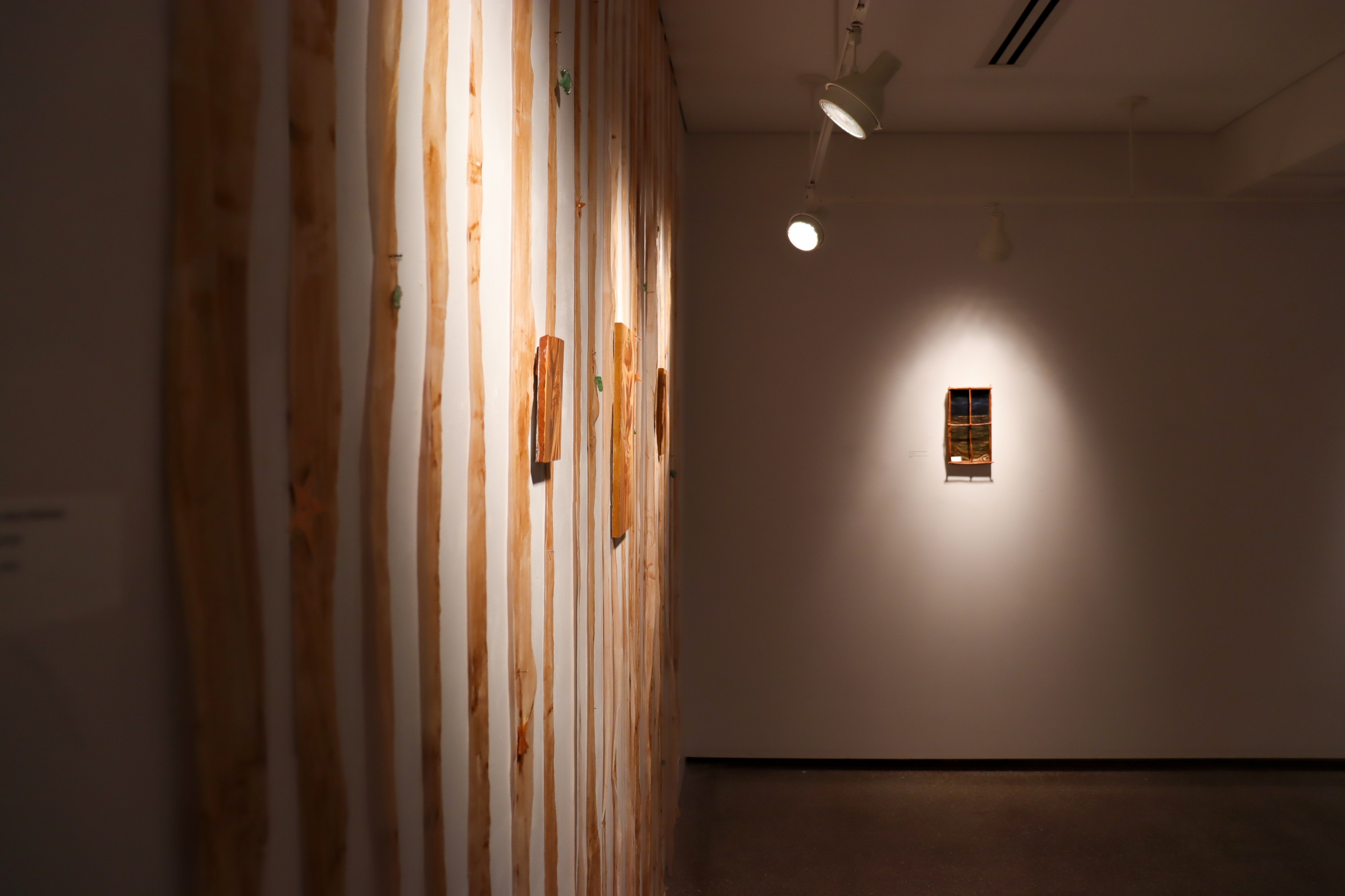 Angled view down the gallery showing the fabric-strip installation along one wall and a small framed work spotlighted on the far wall.