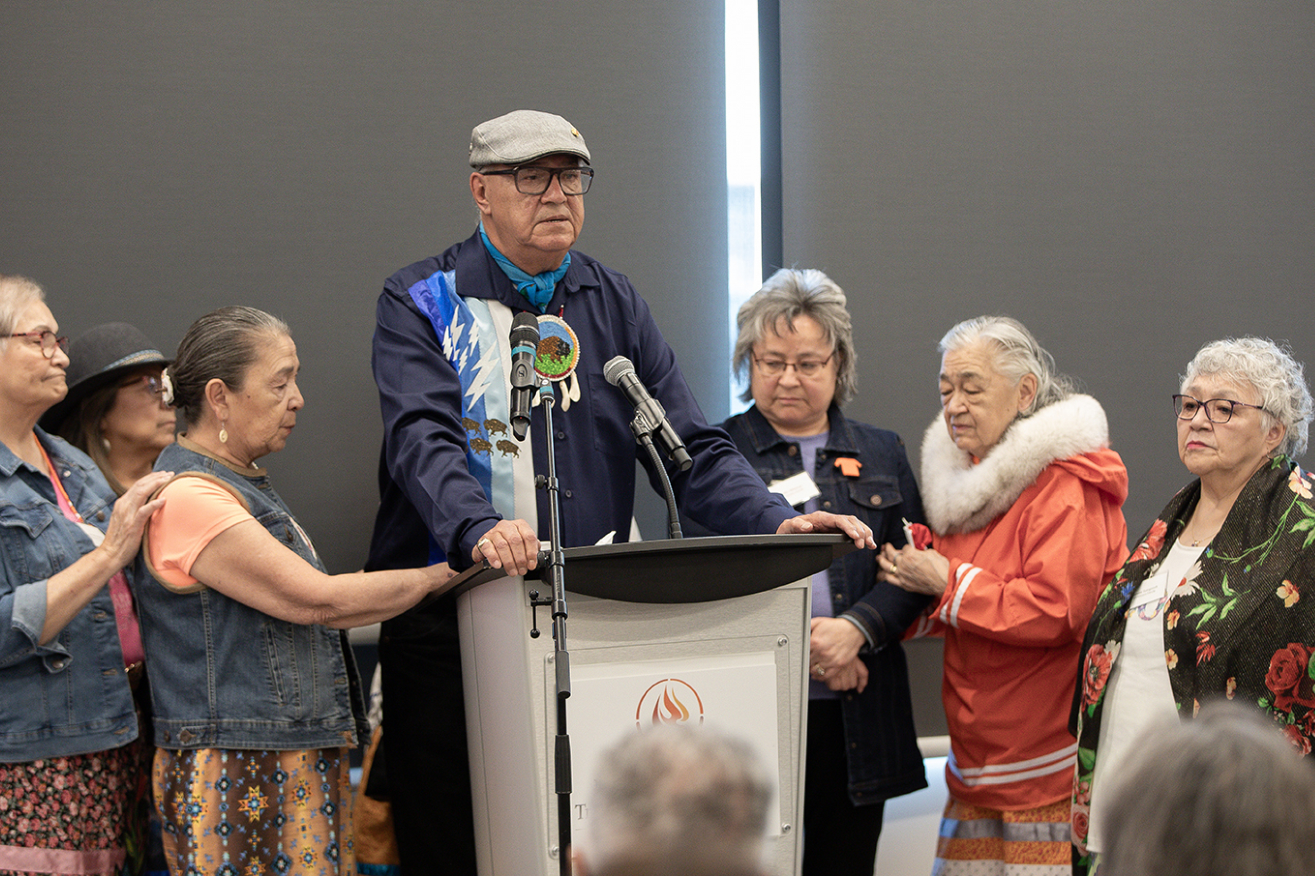 Survivor Eugene Arcand at a podium surrounded by other Survivors