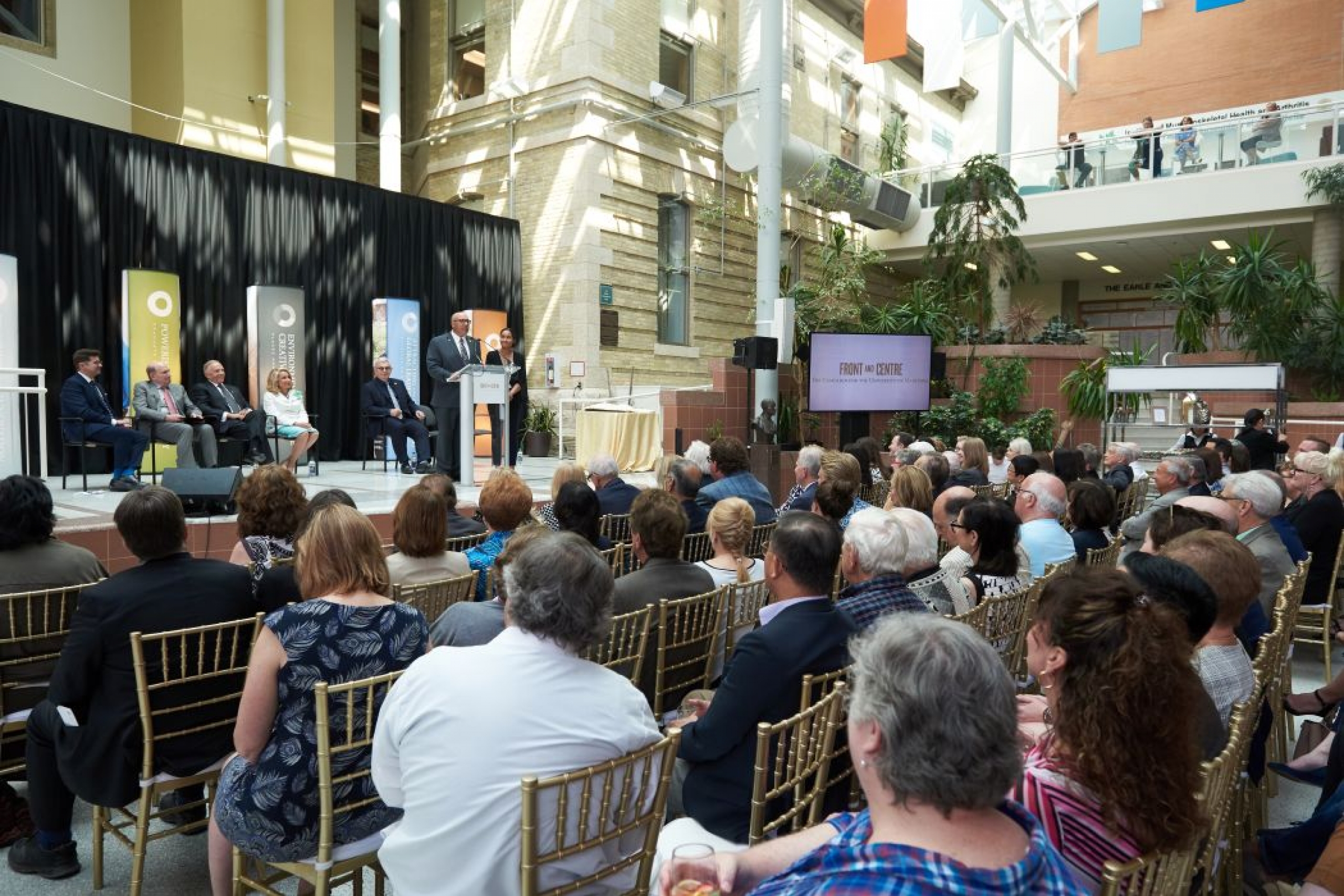 Five people sit in chairs on a stage. Two people stand on stage, one of them speaks at a lectern before a seated audience. 
