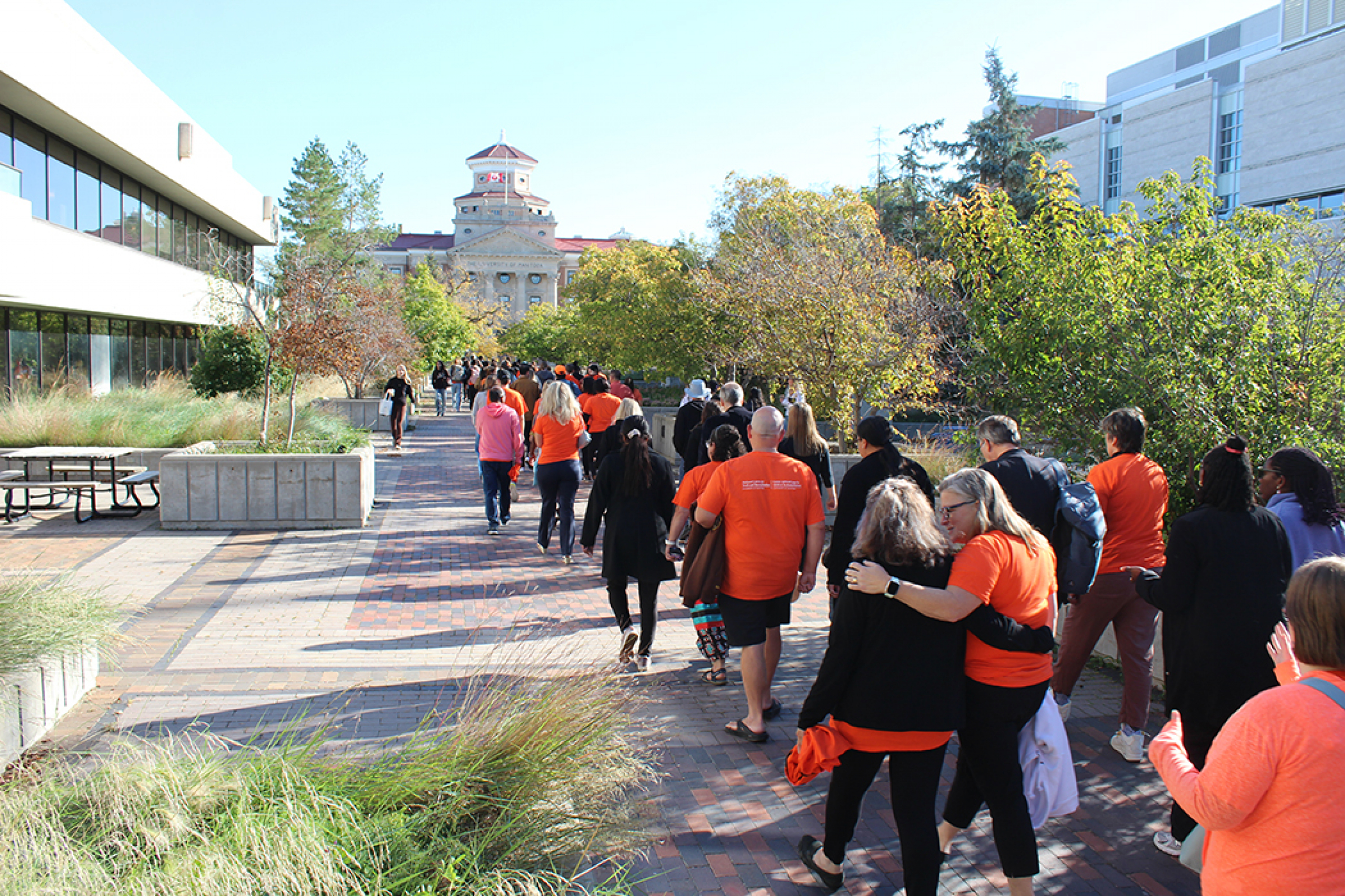 A crowd of people, mostly wearing orange, walk toward the University of Manitoba administration building.
