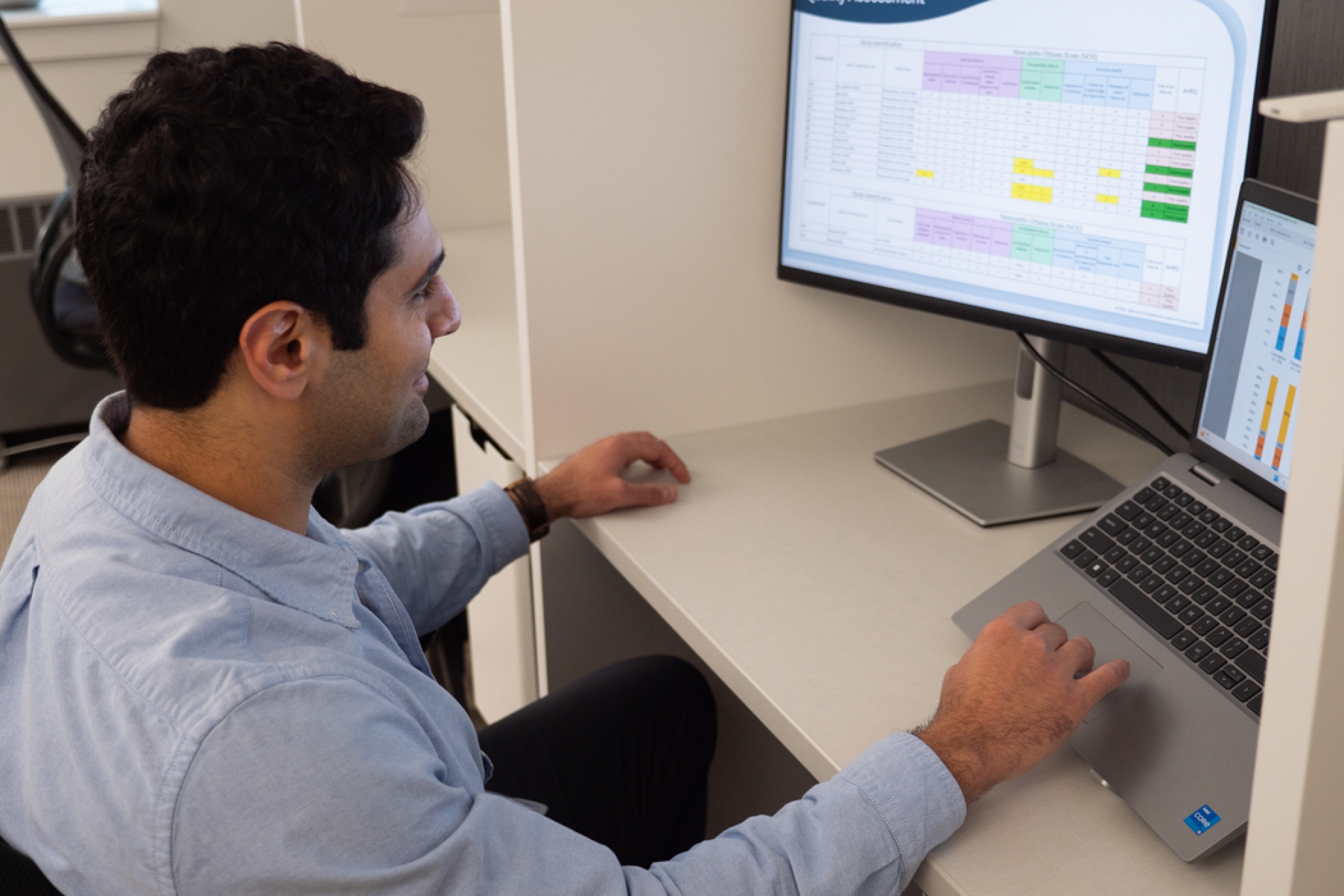Sam Kavosh sits at a desk, looking at a dual-monitor setup. The left screen displays a table titled 'Quality Assessment'.