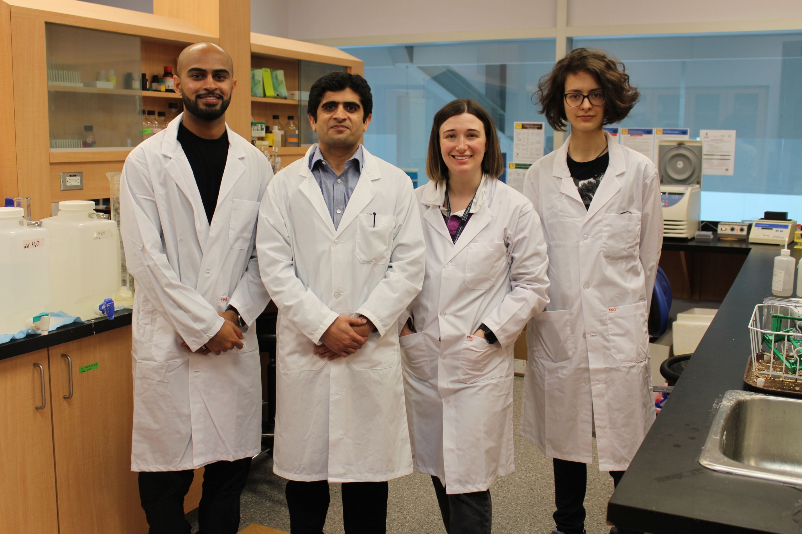 The professor and his grad students pose for a photo in his lab. They are wearing lab coats.