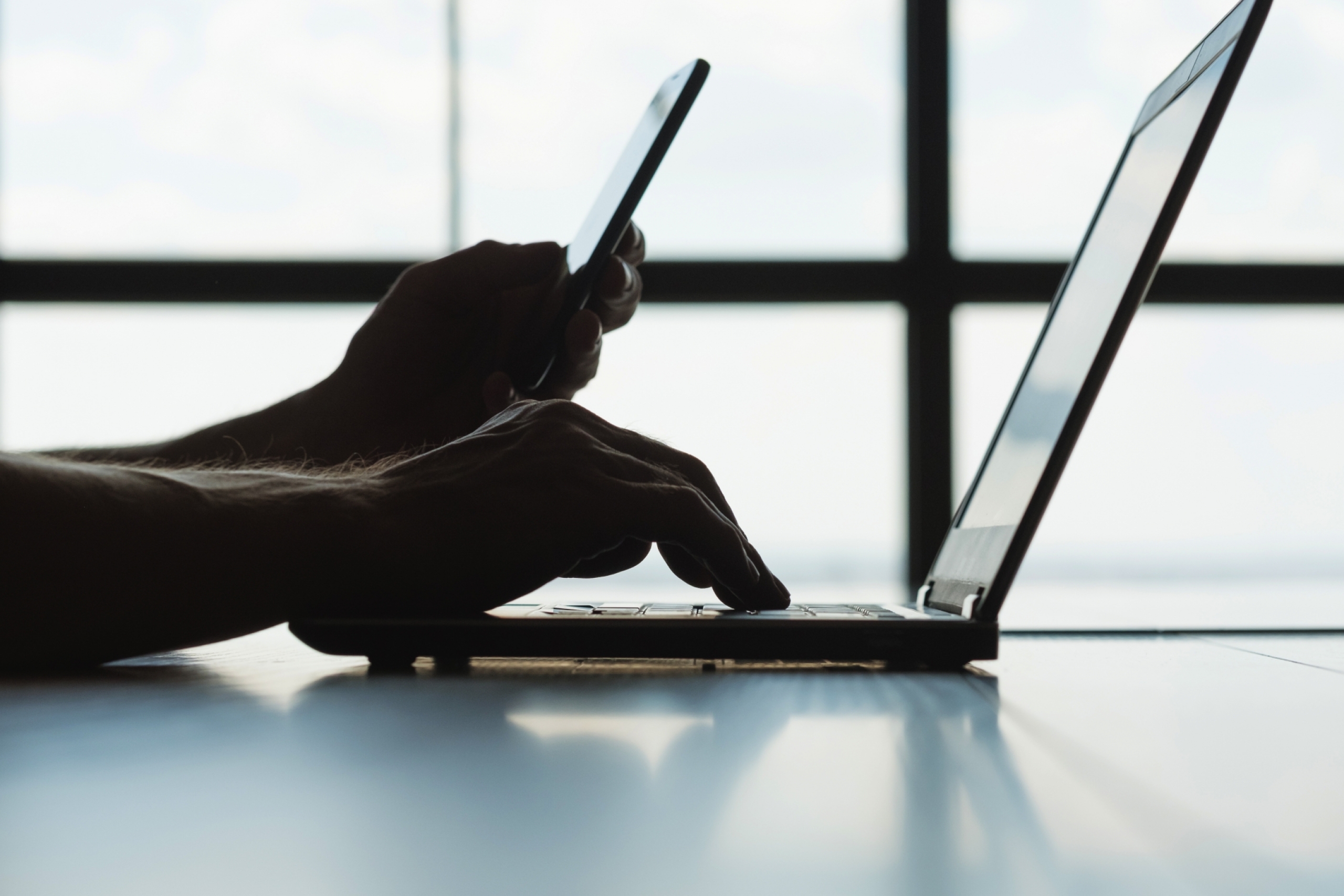 Backlit photo of someone using a phone and a laptop.
