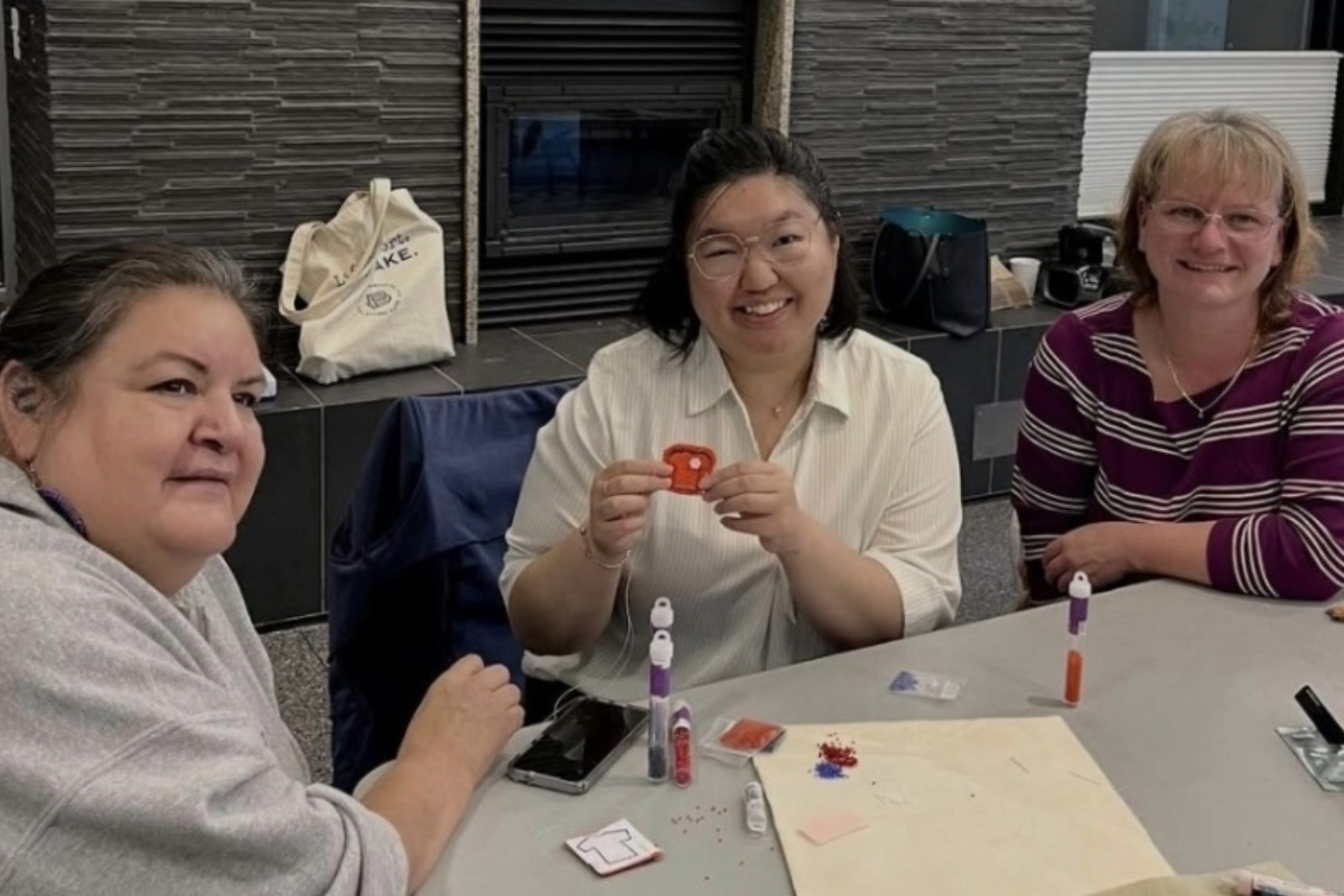Three people pose for a photo, one displaying a tiny beaded orange shirt artwork.