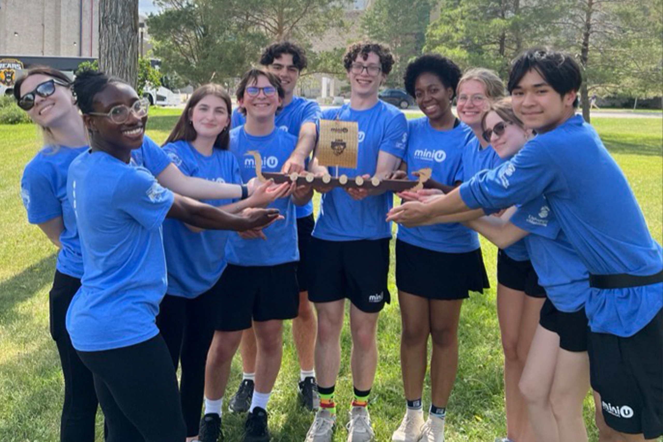 A group of young people in matching blue shirts lean in a semi circle to hold a boat trophy.