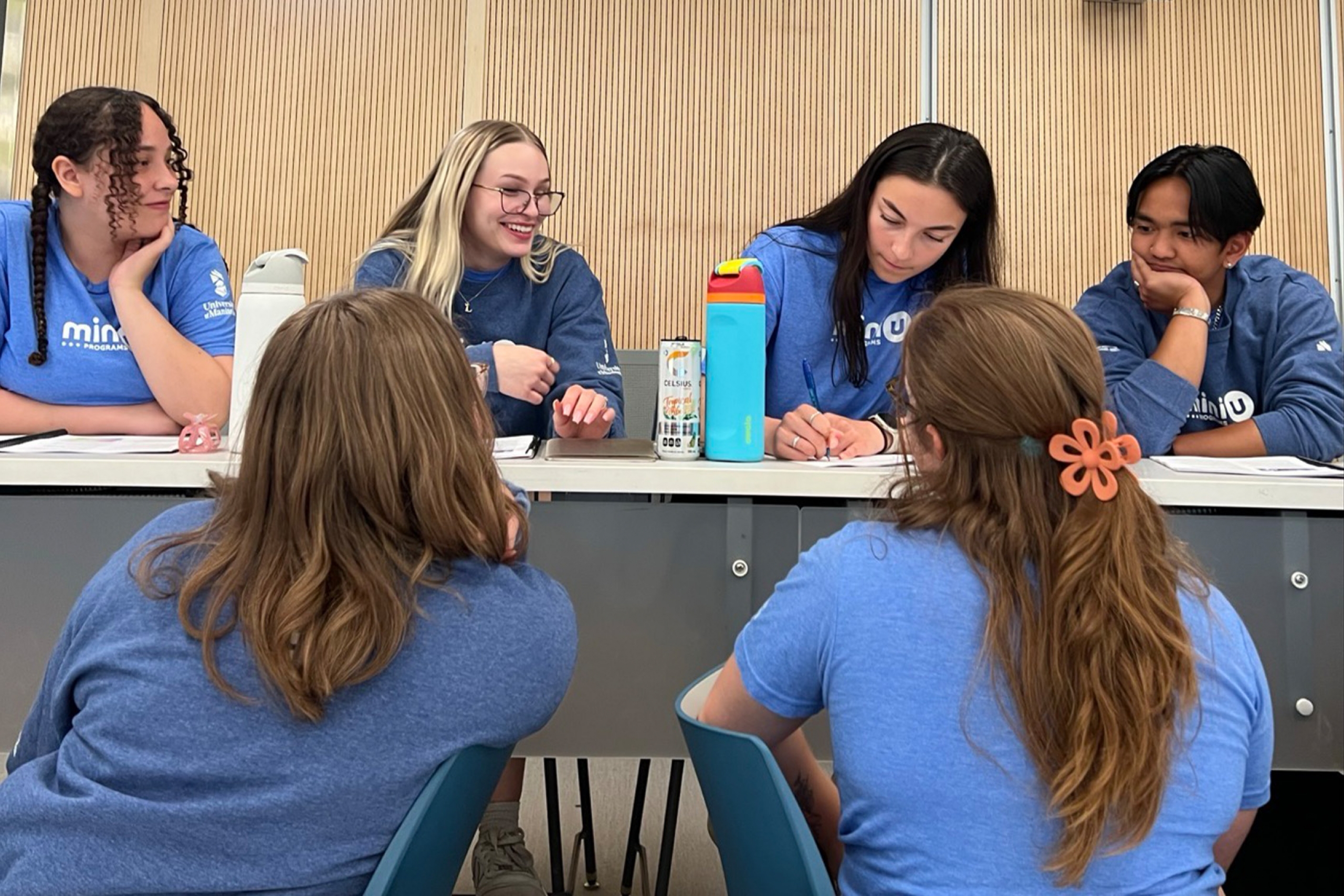 Two rows of young people sit facing each other in a lecture hall. They are wearing matching blue shirts.