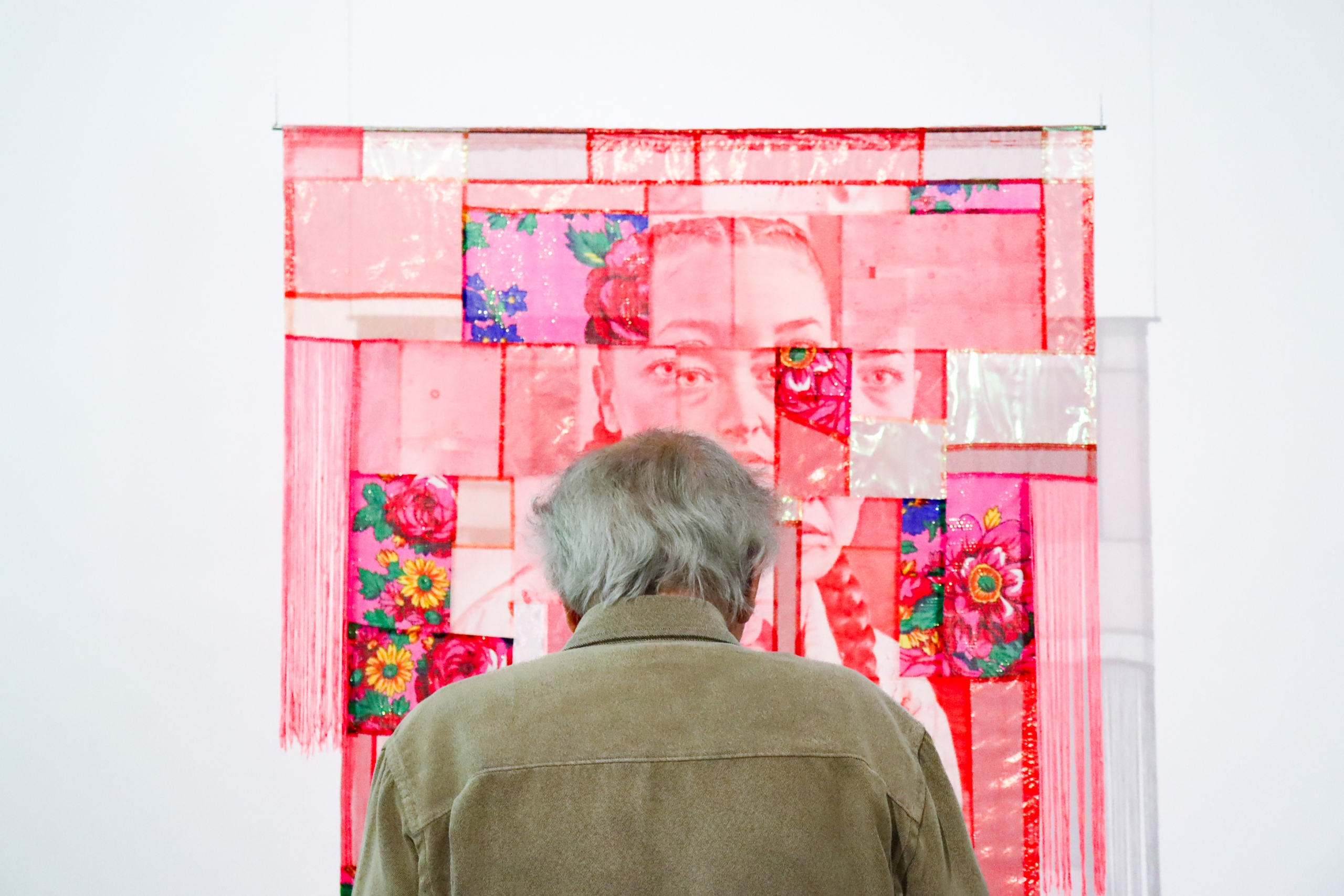 An older visitor stands facing a large red portrait quilt panel with floral blocks and translucent sections.