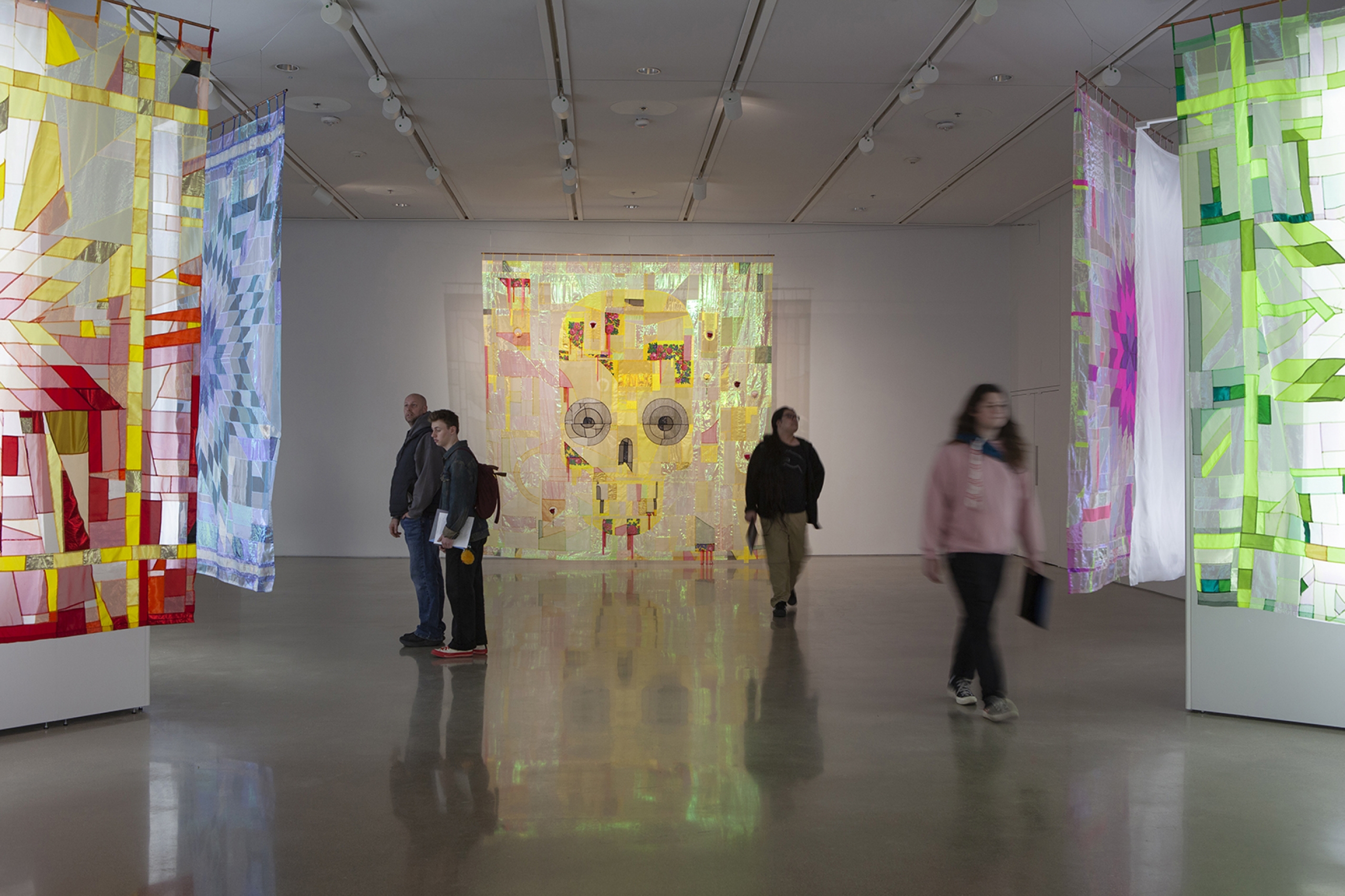 Wide gallery view with visitors walking among suspended textile panels in bright colours and patterns.