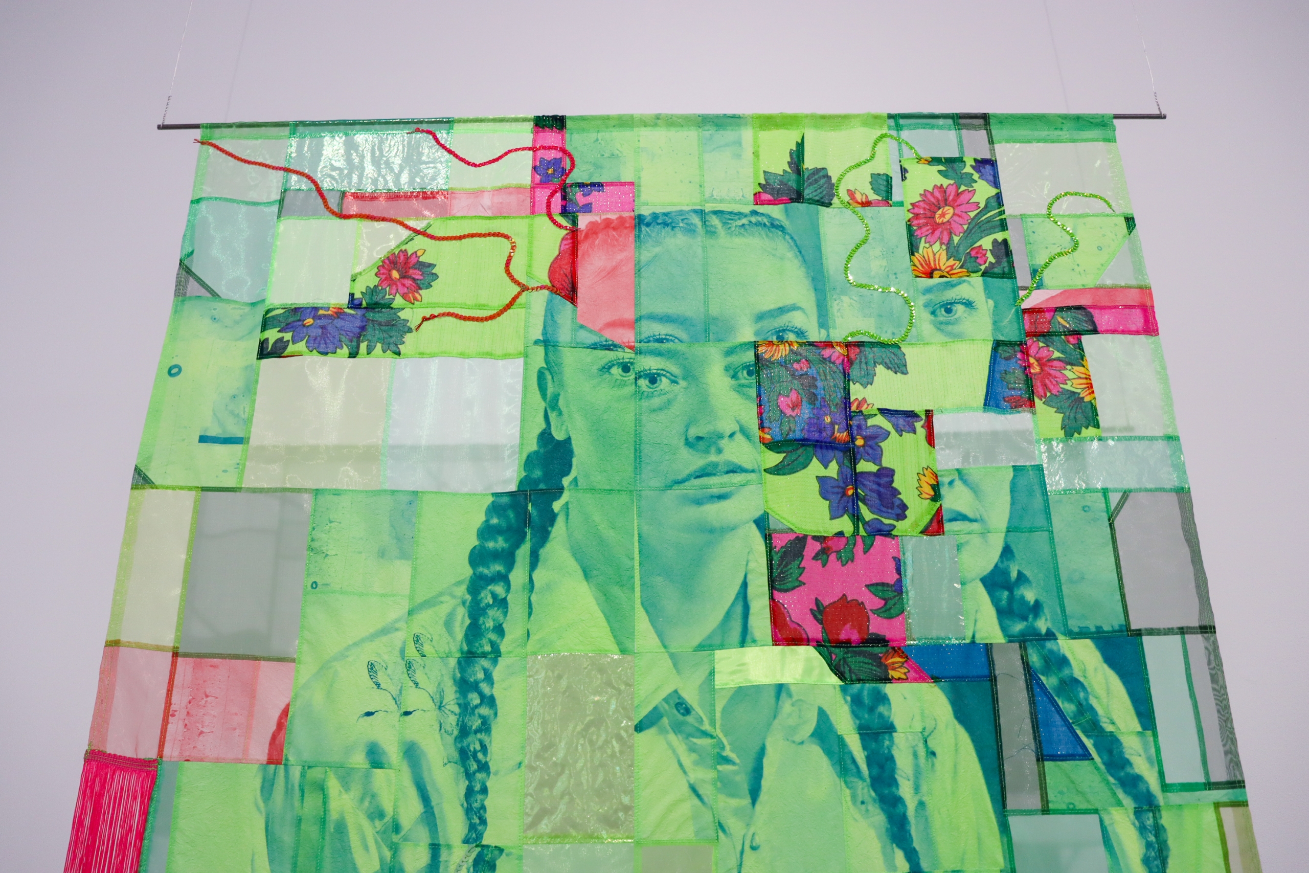 Large green-toned portrait quilt panel with floral patches and braided hair imagery hangs against a white gallery wall.