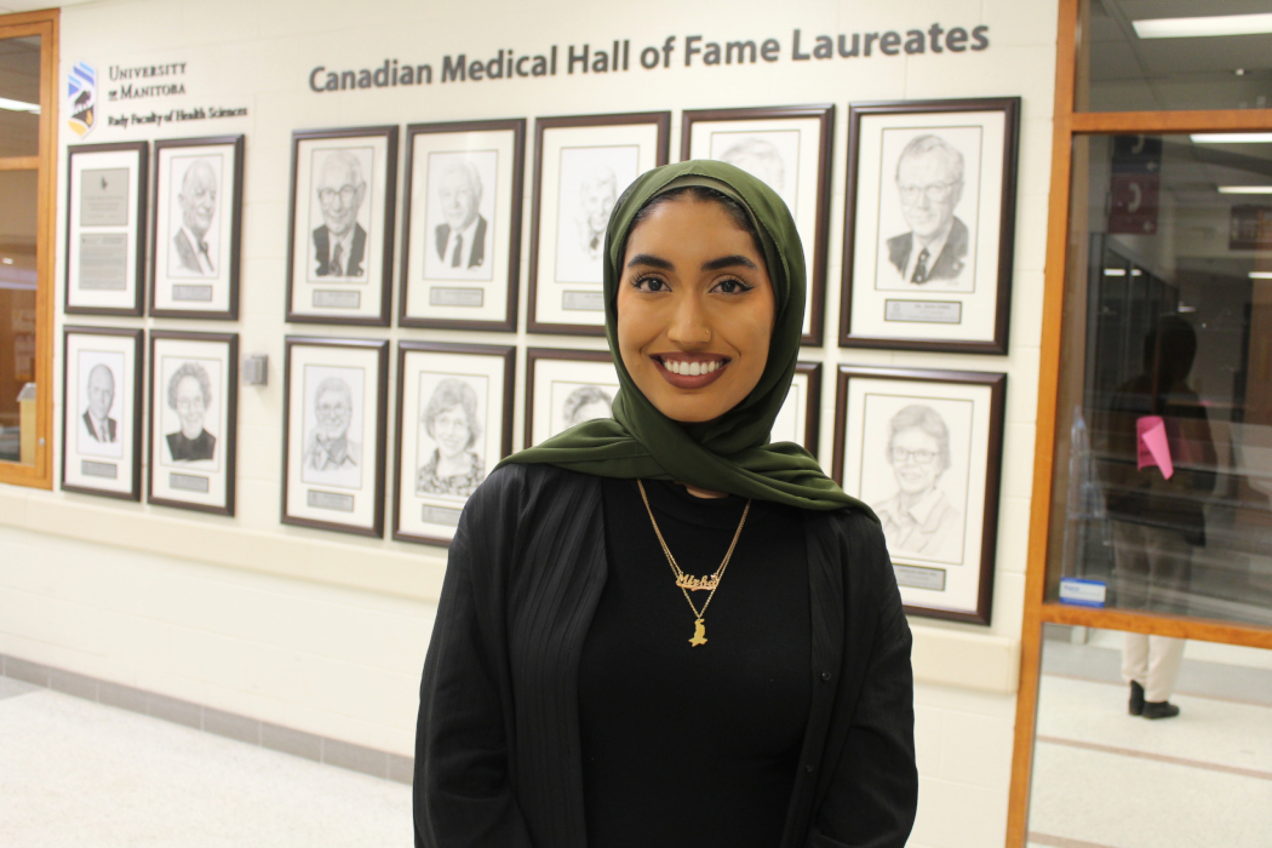 Mirha Zohair poses in front of 13 framed illustrations of Canadian Medical Hall of Fame laureates from UM. Text on wall reads "Canadian Medical Hall of Fame Laureates."