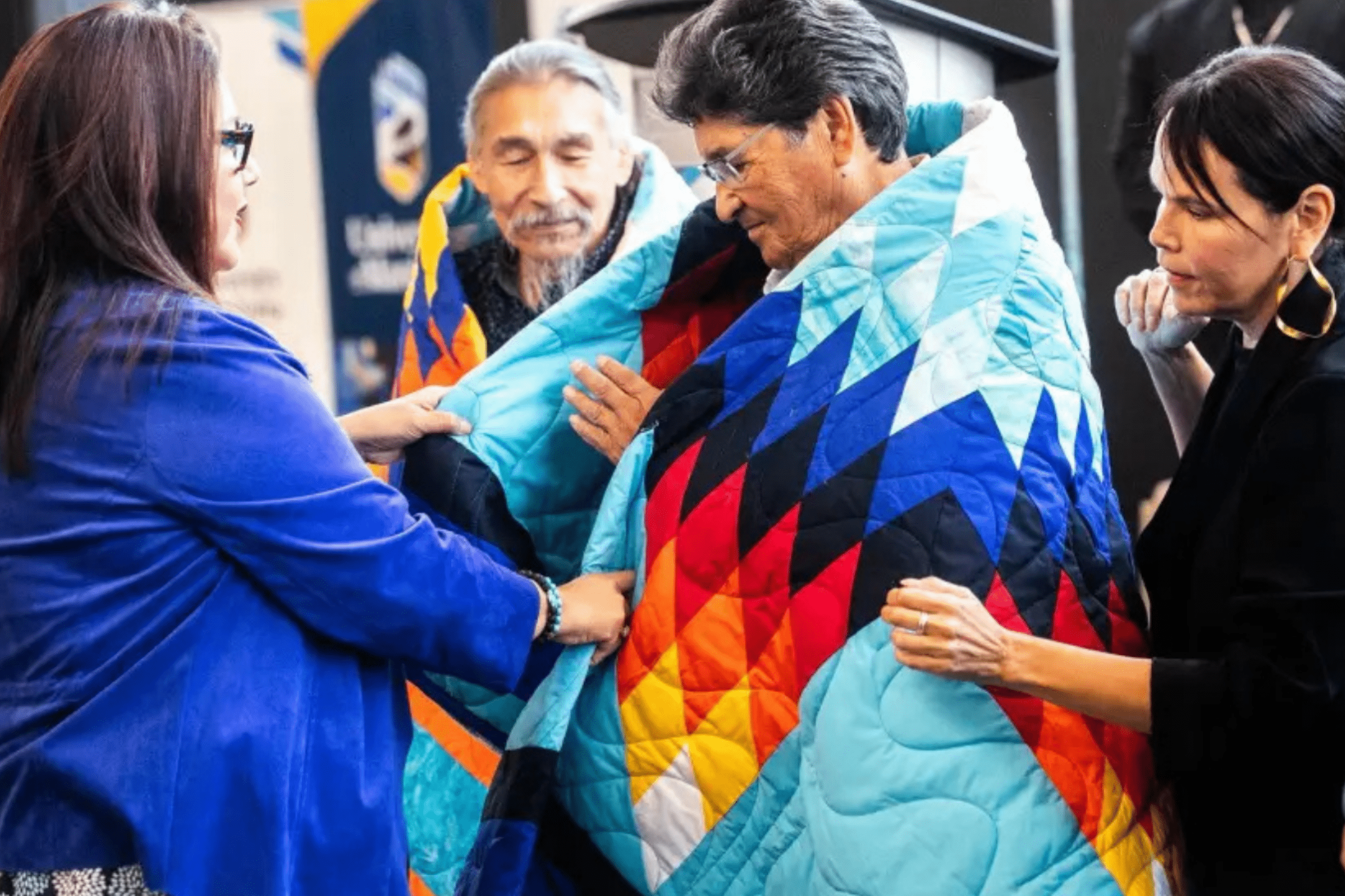 Two Elders being wrapped in colourful star quilts during a blanket ceremony, supported by several community members.