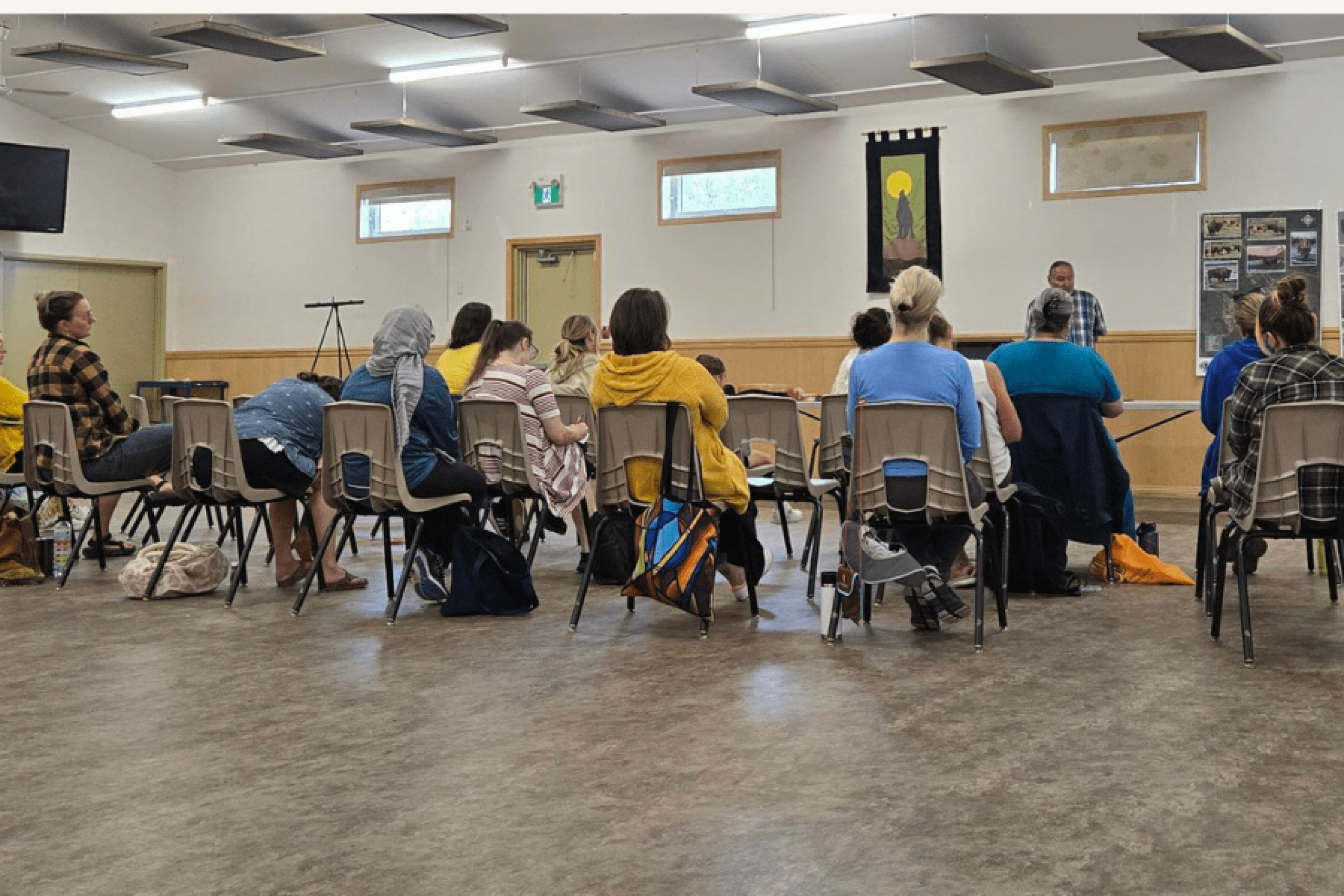 Participants in the Summer Institute sit in a circle during a field trip workshop at Brokenhead, listening to a community speaker at the front of the hall.