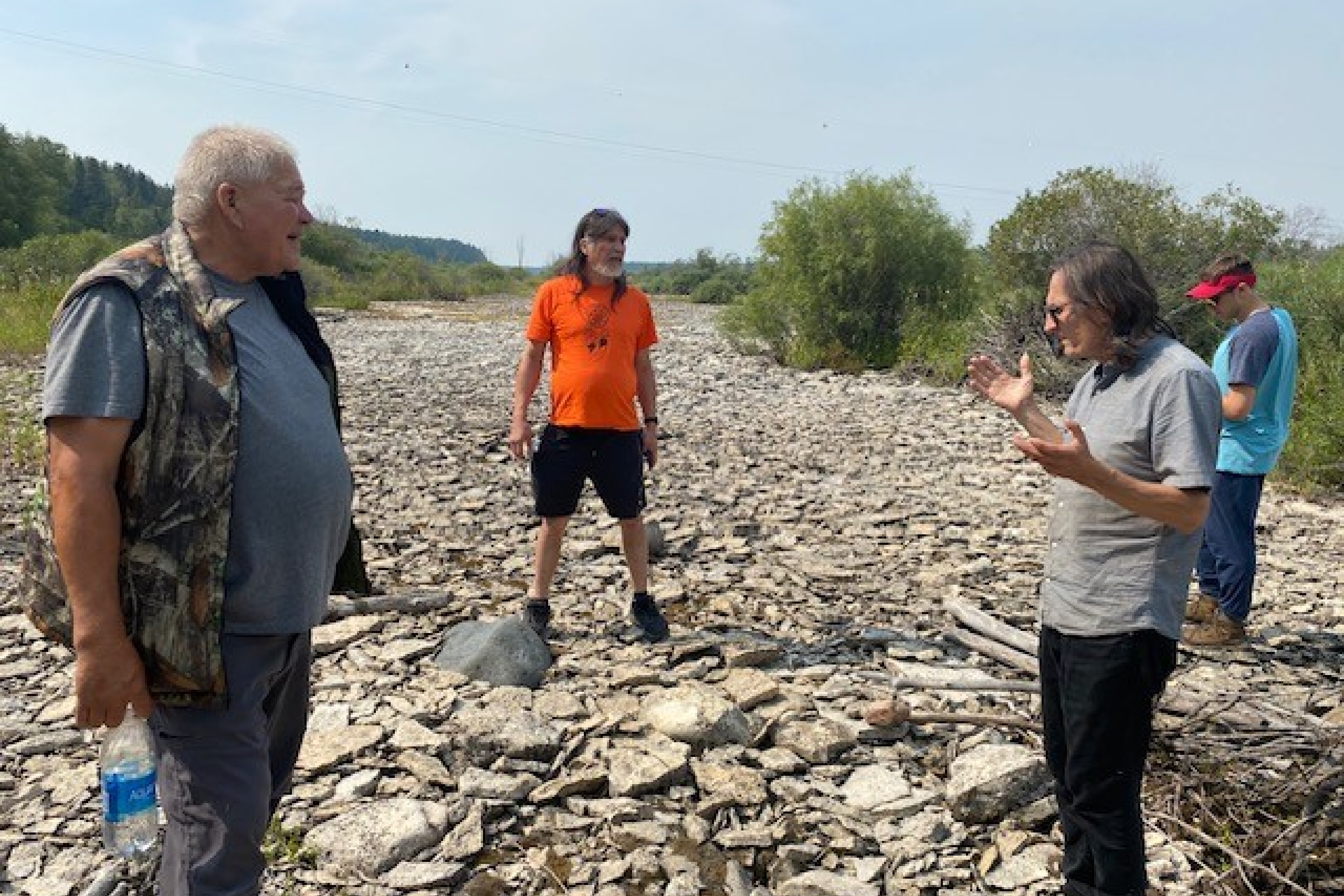 three individuals stand talking together on a dry rocky riverbed.