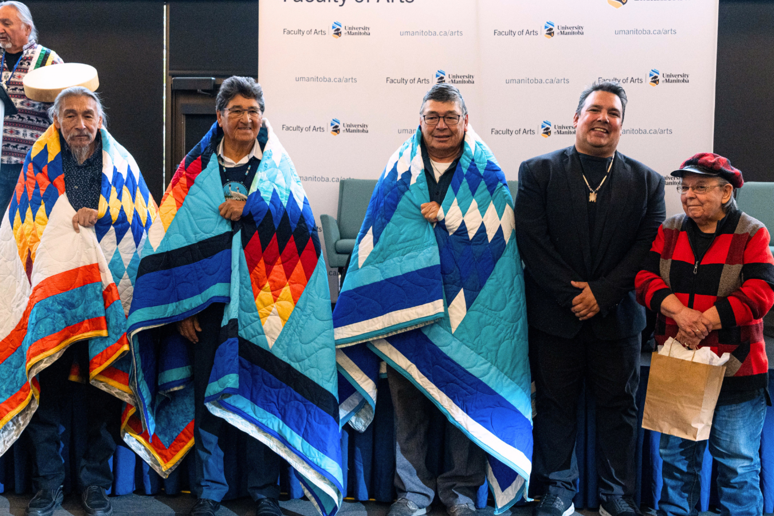 A group of Elders standing together wearing vibrant star quilts during a recognition ceremony, accompanied by additional community members.