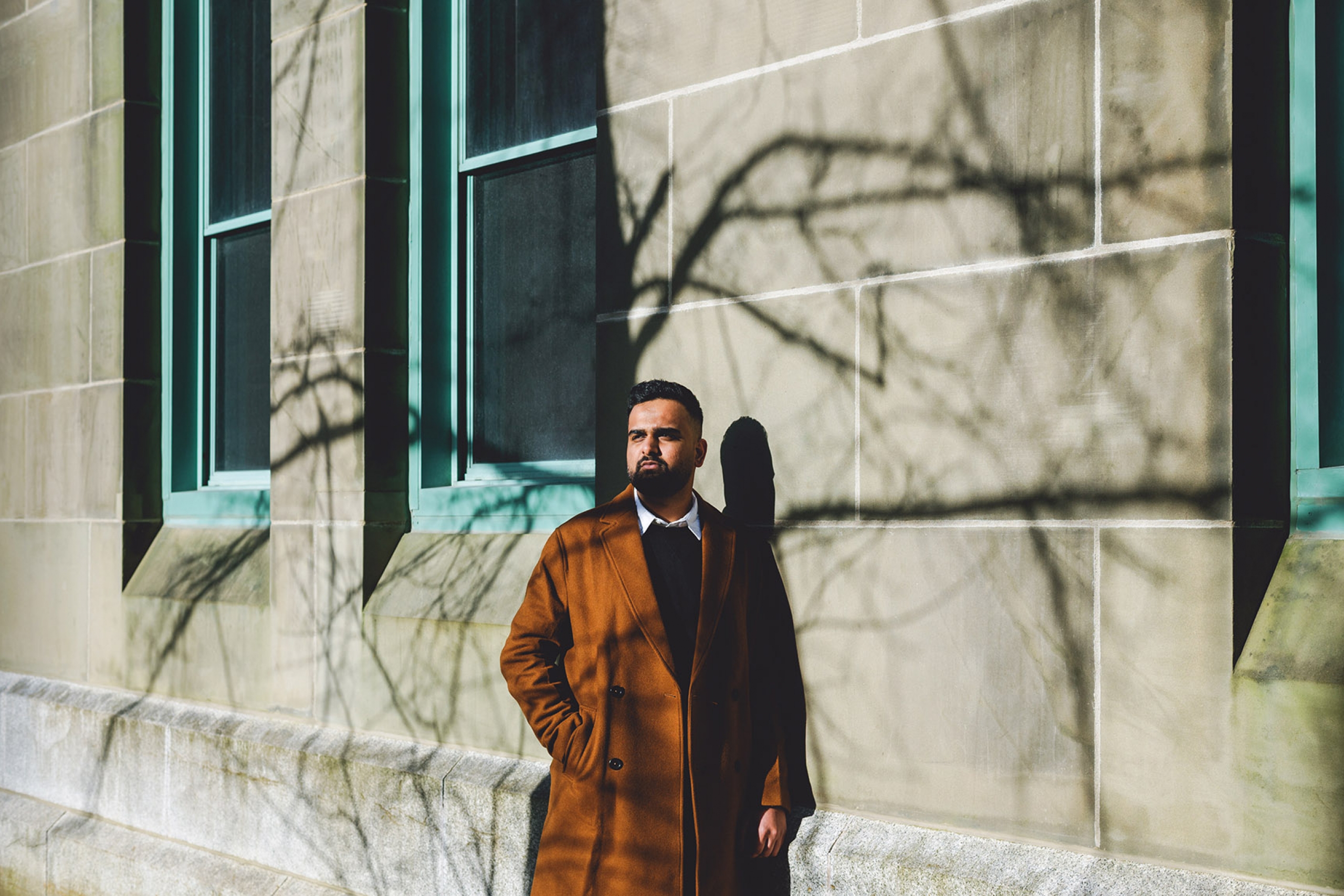 Nabil Iqbal stands in the sunshine, leaning against a building, with the shadow of tree branches cast on the wall behind him.