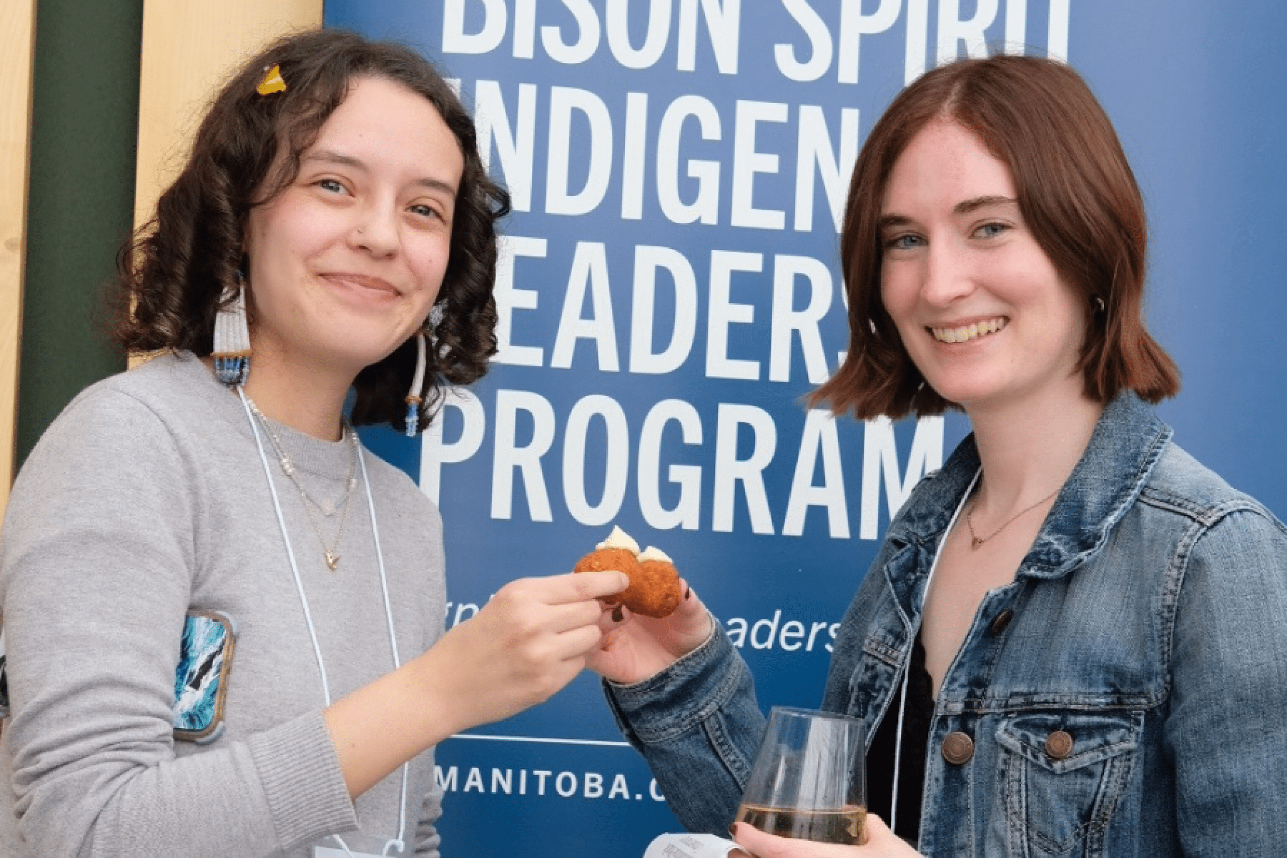 Two students smile and share food in front of a Bison Spirit Indigenous Leadership Program banner.