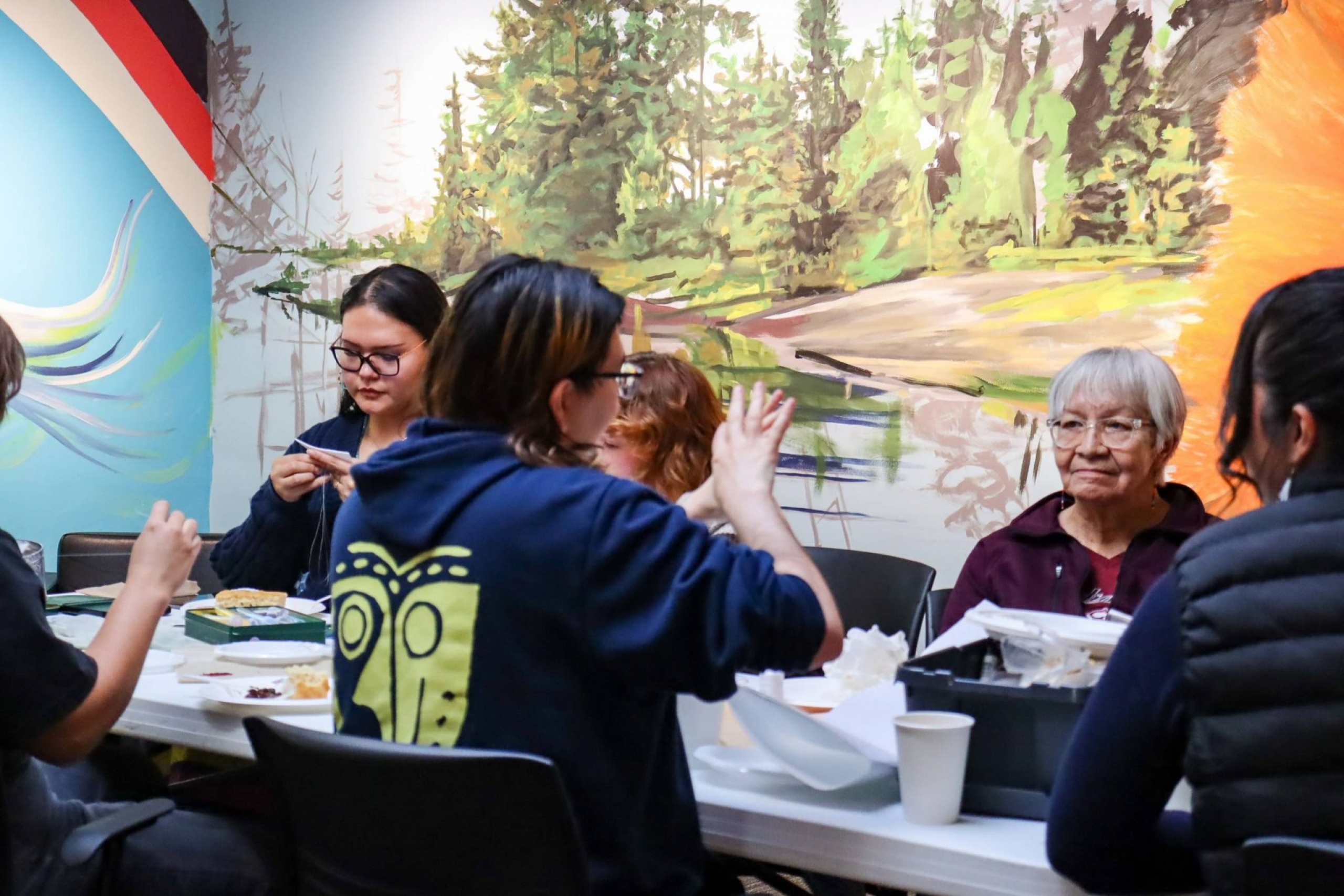 Students and an Elder seated together at a table in front of the mural, chatting and working with craft materials.