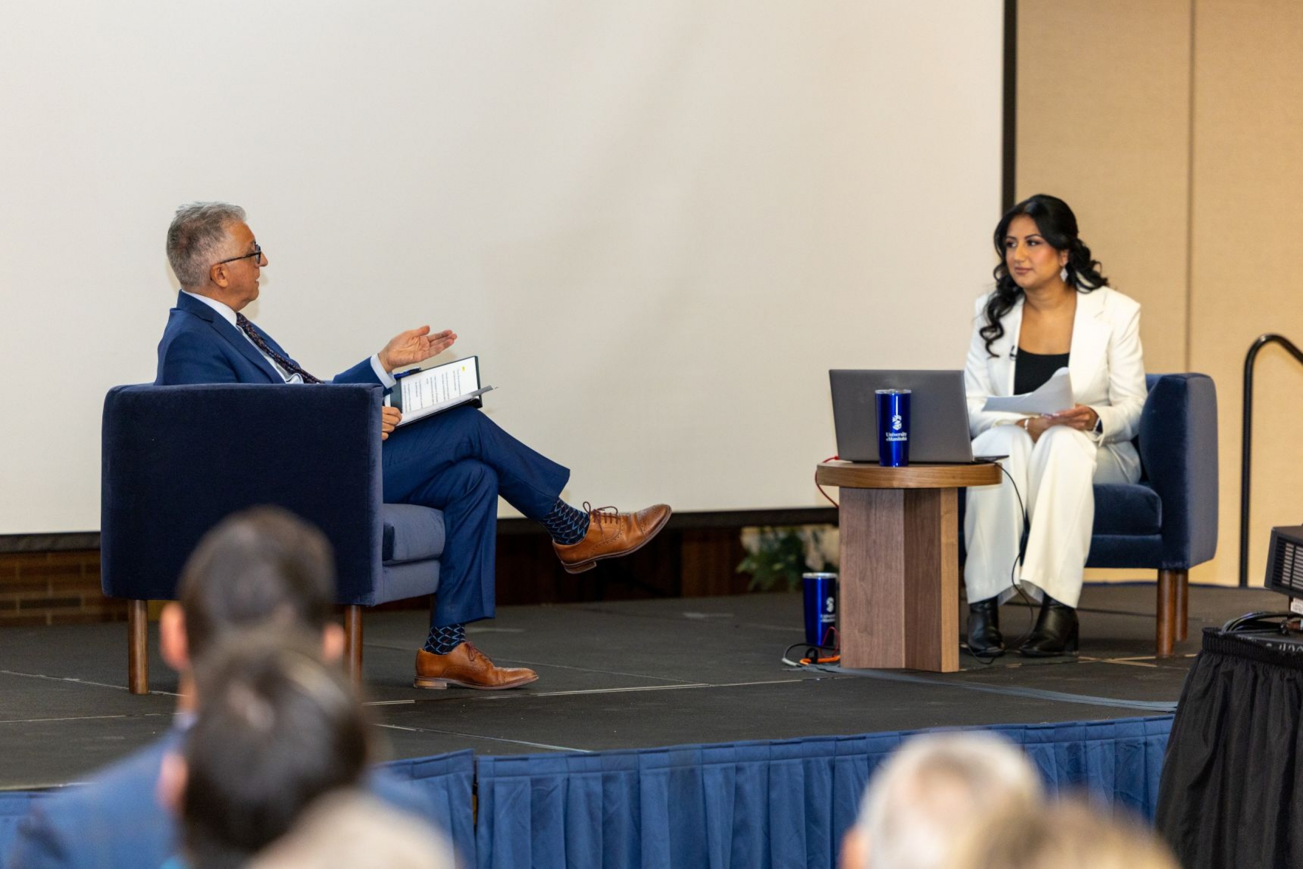 Michael Benarroch and Raman Dhaliwal sit on a stage in an event room.