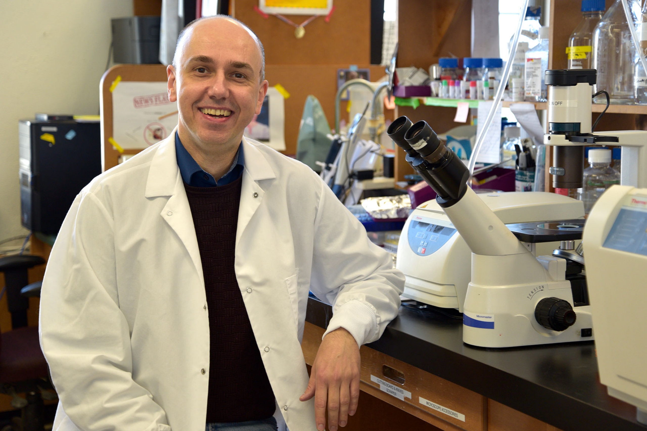 Dr. Peter Pelka wearing a lab coat beside a microscope smiling at the camera.