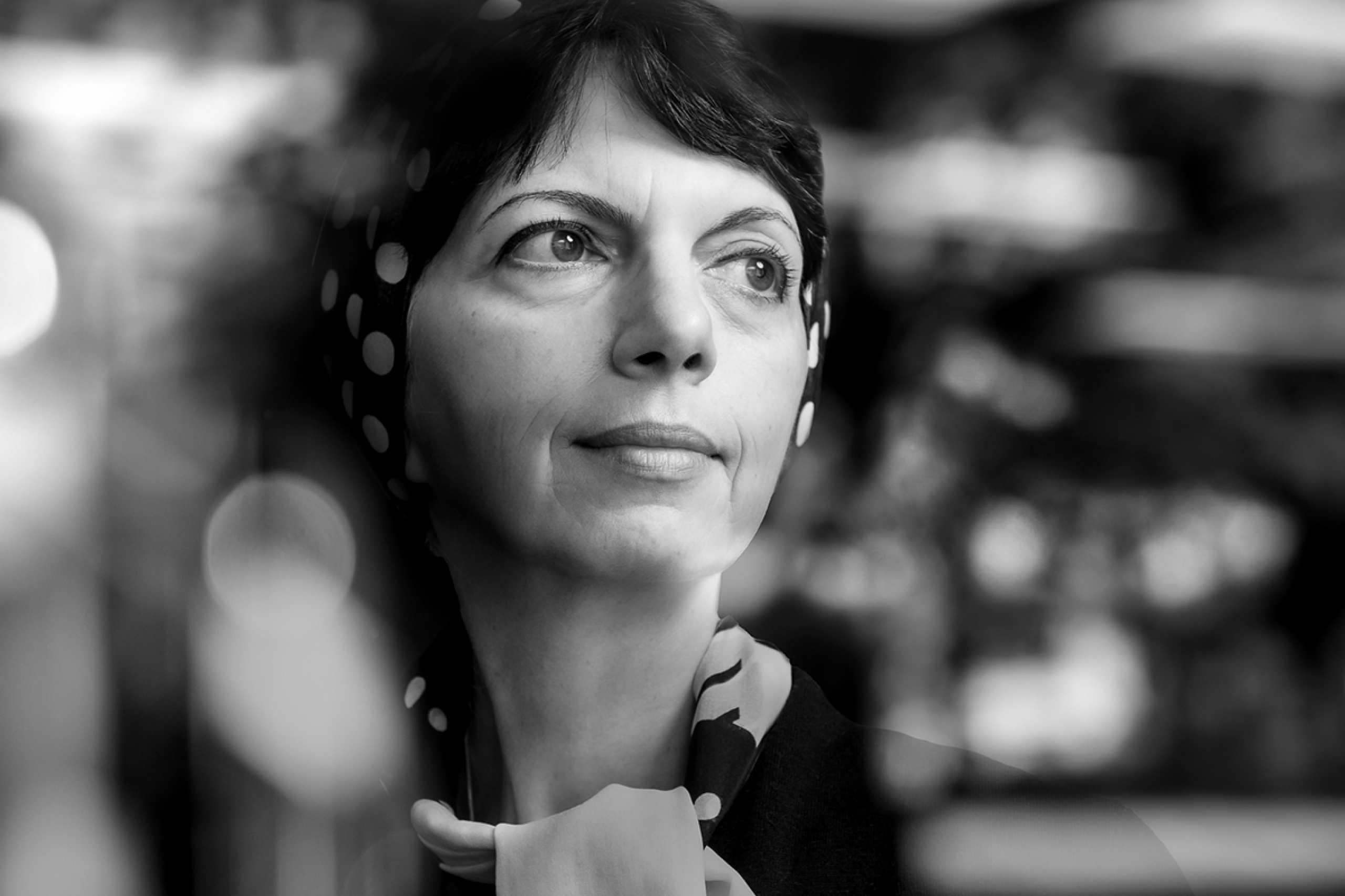 Zahra Moussavi looks out a window with a pensive look on her face. Blurred trees can be seen in the window's reflection.