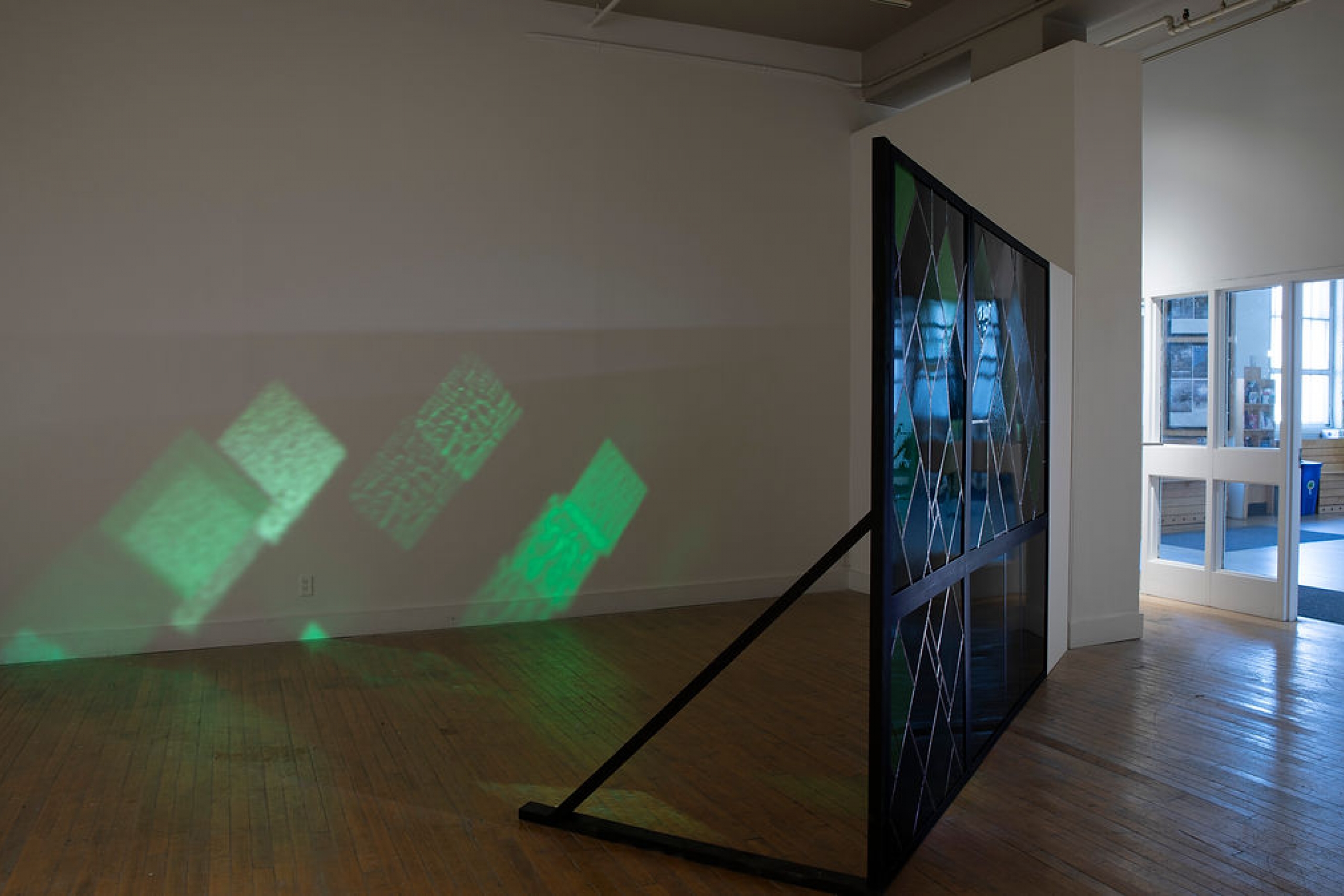 A metal lattice glass sculpture stands on a concrete base in a gallery space, with a small brick-and-glass wall piece mounted nearby.