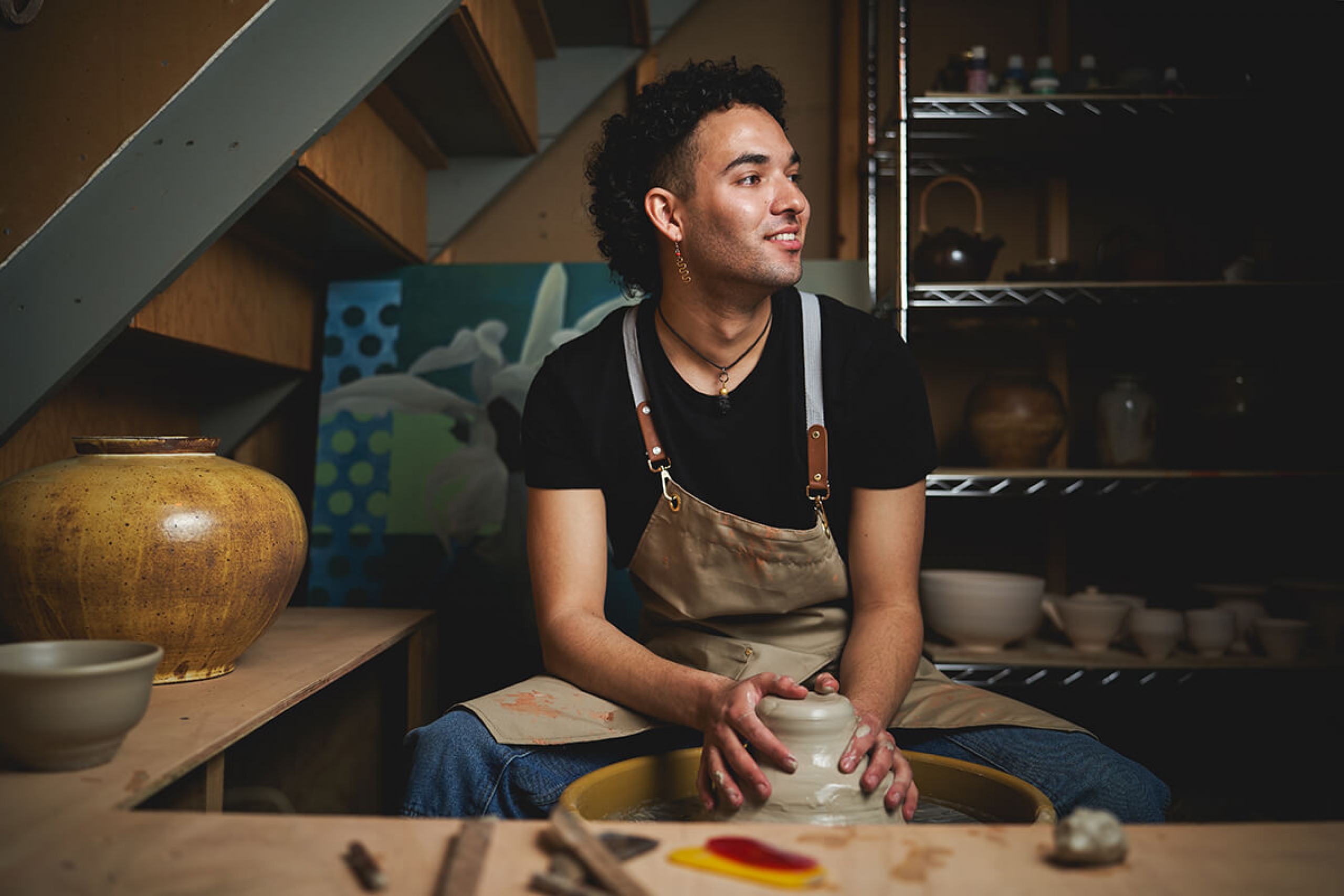 Joel Nichols sits on a stool while working on a ceramic in his basement art studio