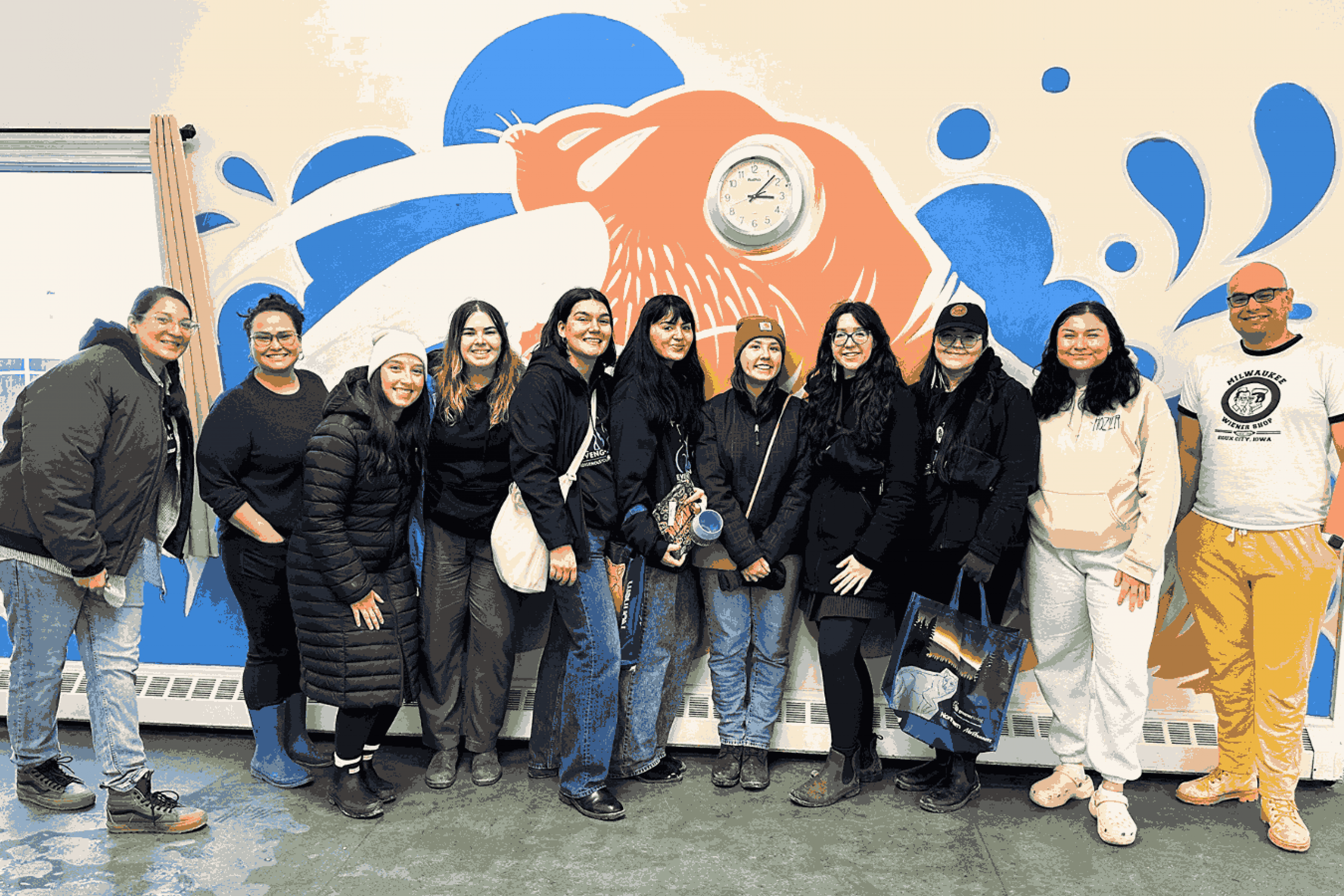A group of people stand together smiling in front of a colorful mural featuring an orange and blue animal design.