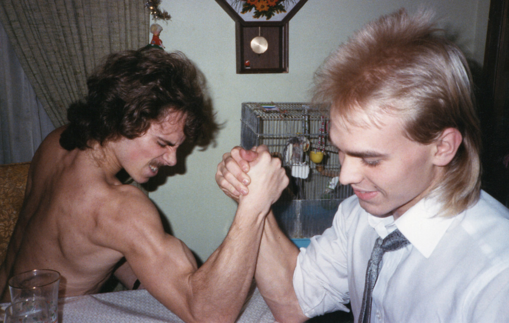 Two guys in the 80s arm wrestle. One has a mullet and is shirtless. The other has a mullet and is wearing a shirt and tie. They seem to be in a kitchen,. probably drunk, and they have a pet budgie in the background