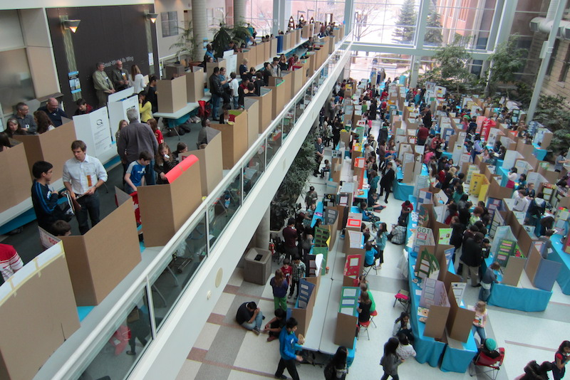 an overhead shot of a science fair, packed aisles and packed tables. one kid totally got his parents to help, you can tell