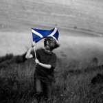 a black and white photo of a boy running through a field carrying a Scottish flag, which is coloured