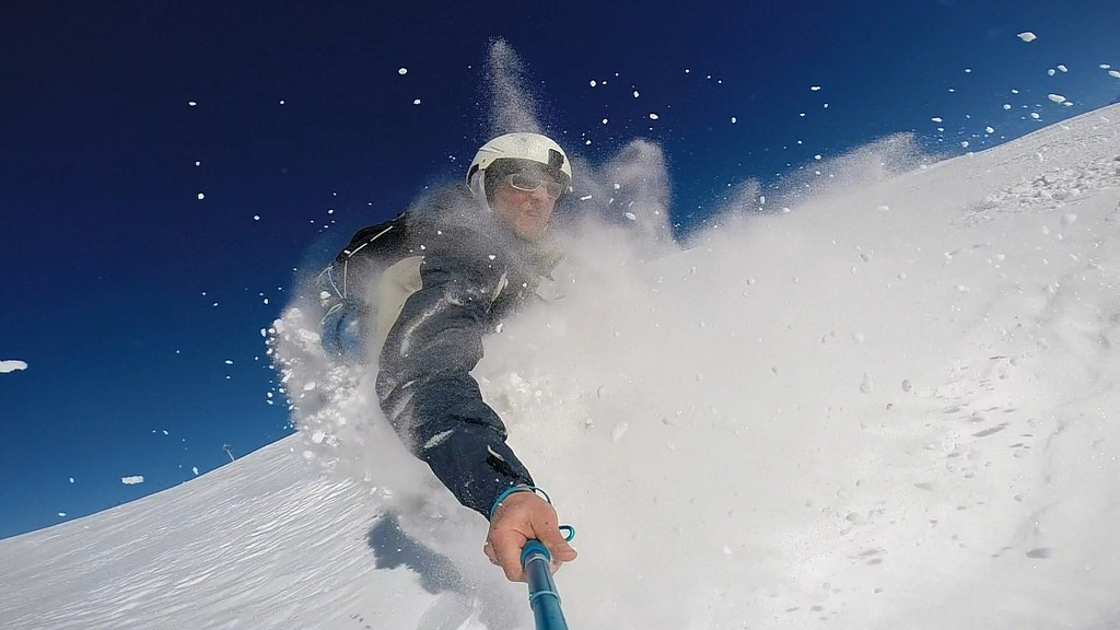 a deep blue sky behind a man snowboarding and kicking up a lot of fresh snow powder into his face