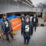 Four students stand outside their makeshift home made of cardboard and tarps