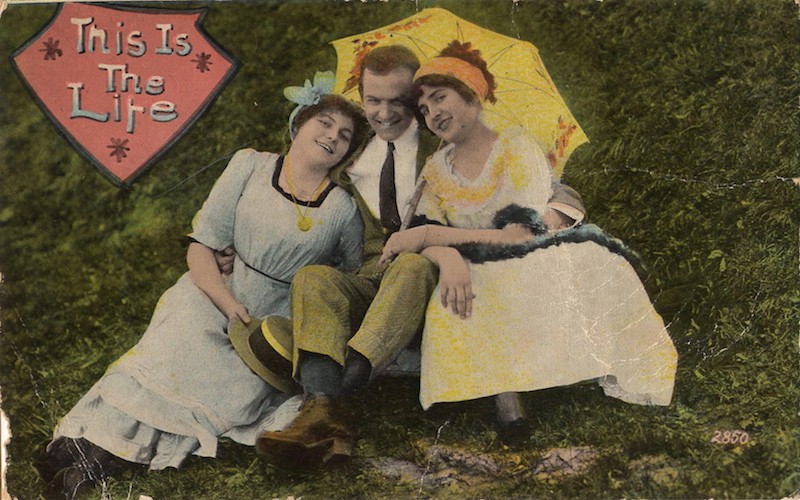A hand tinted postcard from about 1910 shows a man between two women // Wikimedia