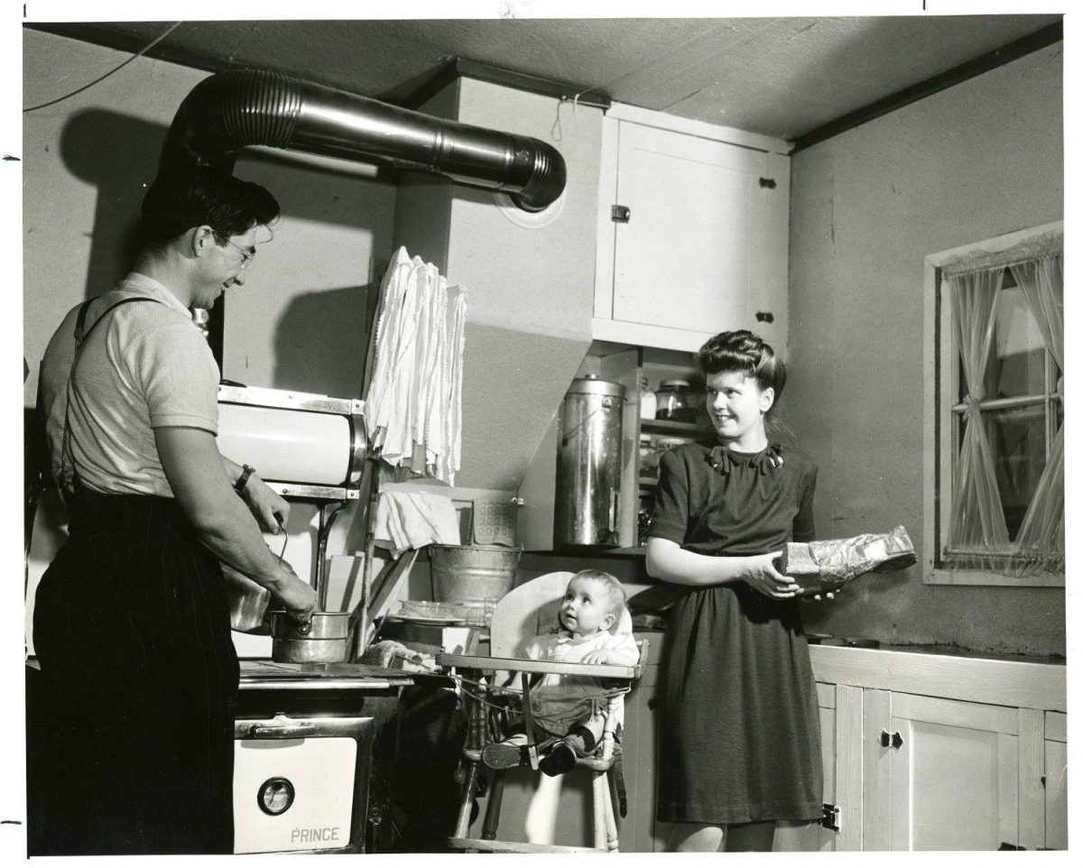 A photograph of a family inside the kitchen of a home in Veterans' Village at the University of Manitoba Campus. A man is standing at the stove with a pot, an infant is in a high chair, and a woman appears to be holding a loaf of bread and a bag in her hands. Photograph is dated as having been taken around 1948 by the National Film Board of Canada. Source: University Relations & Information Office fonds