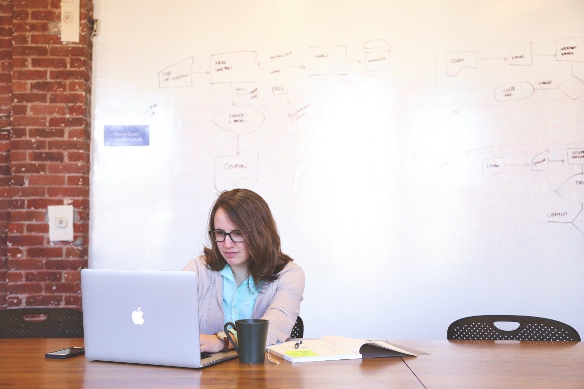 A woman works alone in a board room at a computer in front of a white board