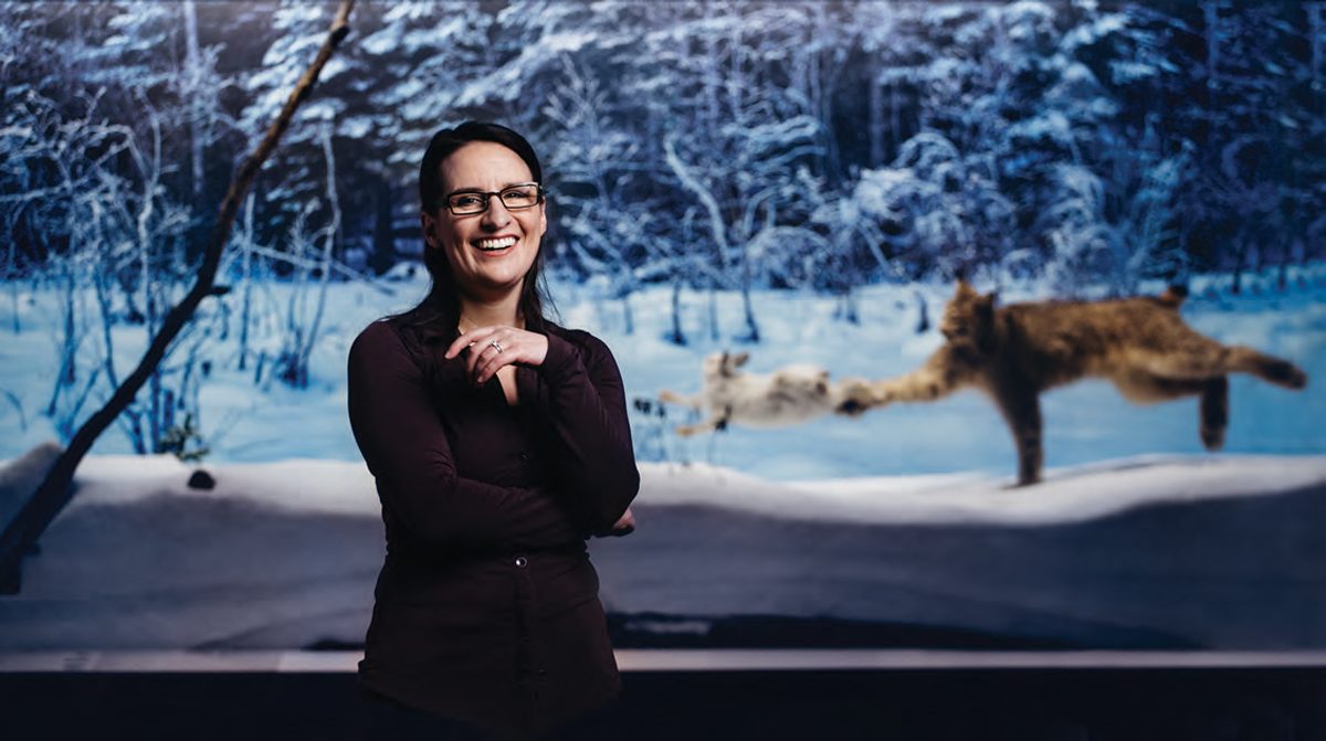 Christa Burstahler stands in front of a diorama of a lynx hunting in winter.