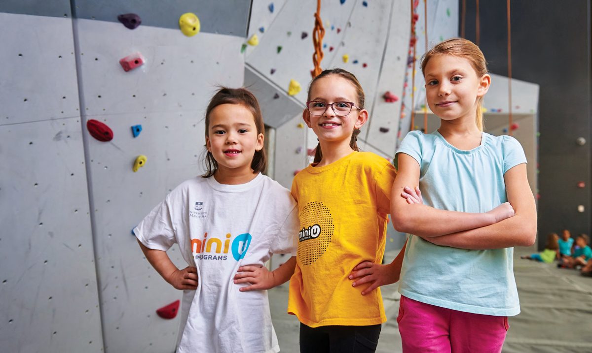 Three girls stand in front of the rock climbing wall at Mini U.