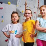 Three girls stand in front of the rock climbing wall at Mini U.