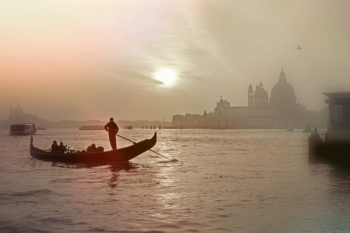 A person paddles standing up in a gondola while the sun shines through haze over Venice.