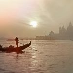 A person paddles standing up in a gondola while the sun shines through haze over Venice.