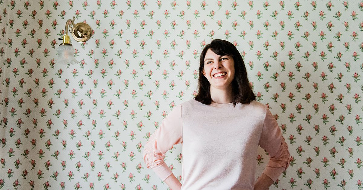 Grace Paizen stands in a pink sweater in front of a floral wallpaper.