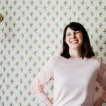 Grace Paizen stands in a pink sweater in front of a floral wallpaper.