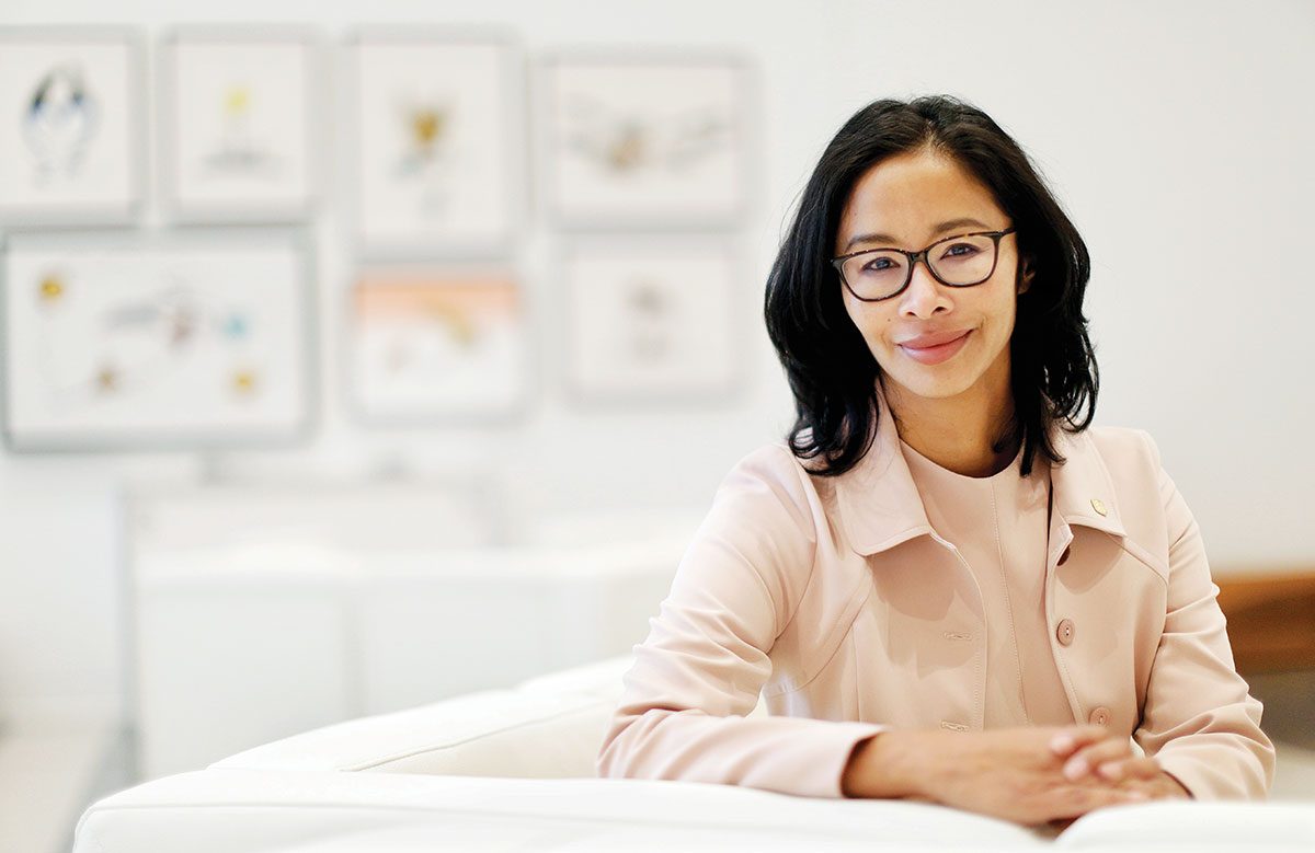 Gigi Osler sits with her hands clasped in front of a wall of framed images.