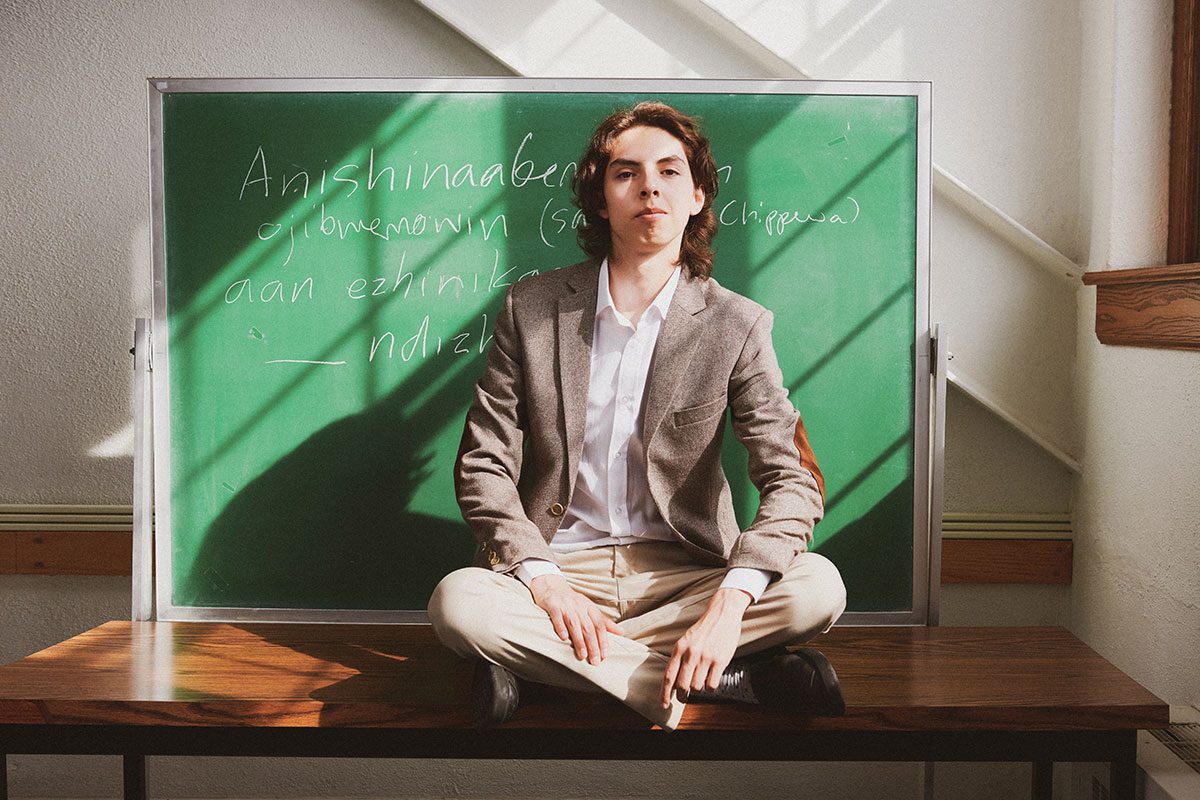 Aandeg Muldrew sits cross-legged on top of a desk in front of a green chalkboard.
